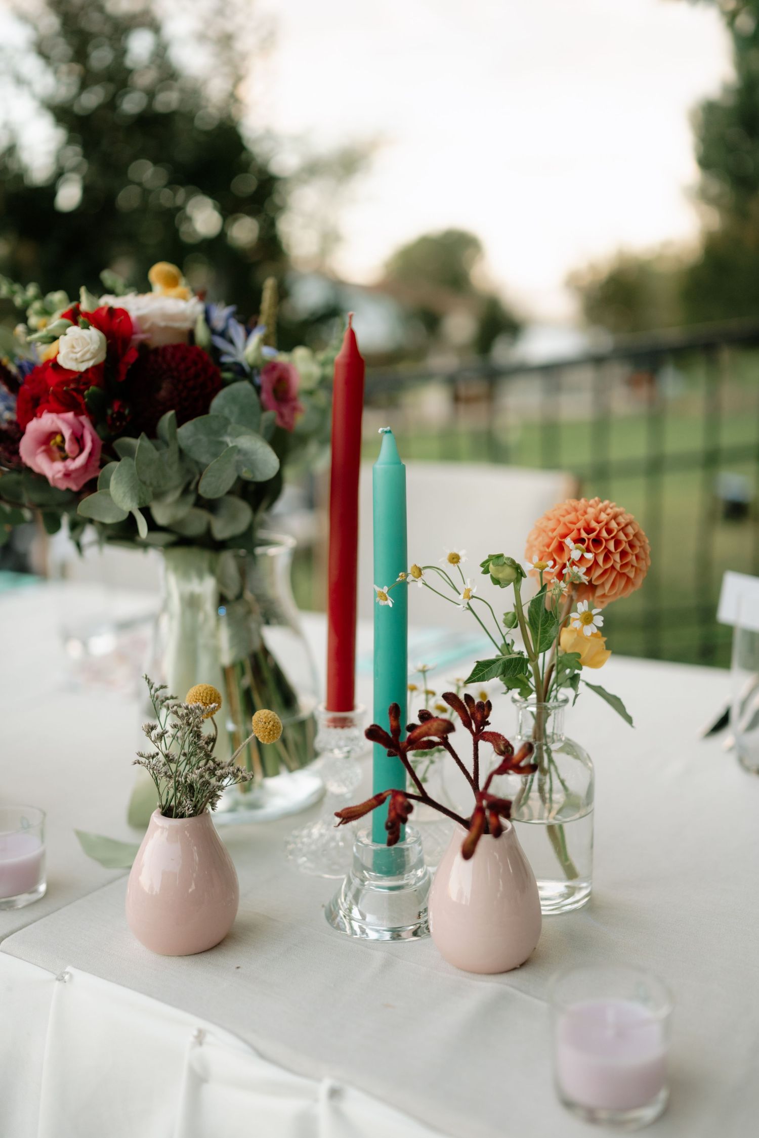 Wedding table decor with pink and red flowers in vases, turquoise and red taper candles on white tablecloth outdoors.