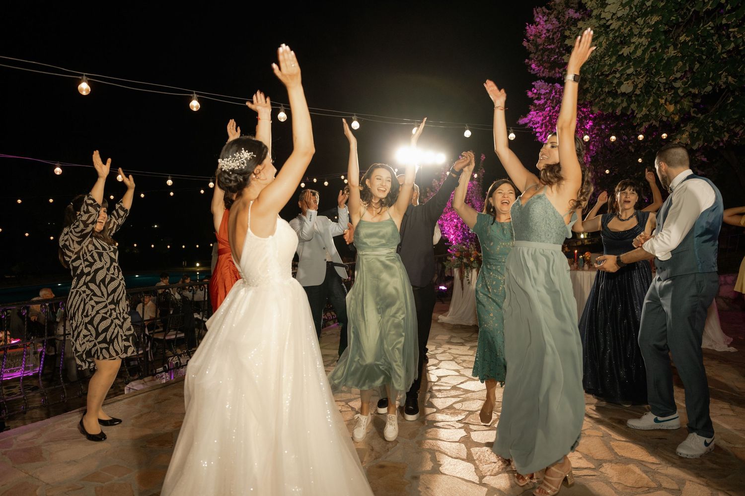 Wedding guests dance and celebrate on an outdoor dance floor under string lights during an evening reception.