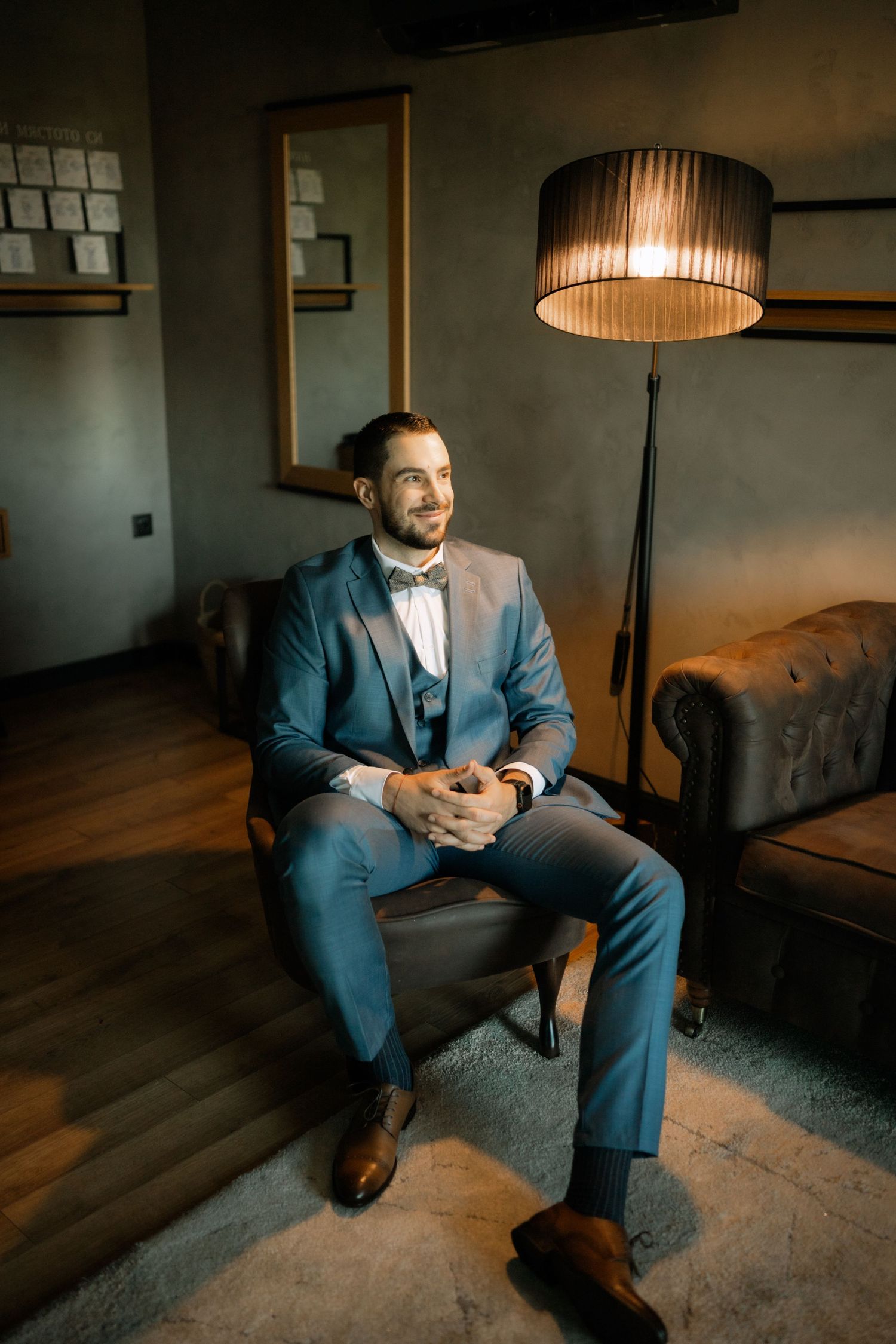 A person in a blue suit sits thoughtfully in a dimly lit room with warm lamp lighting creating a moody atmosphere.