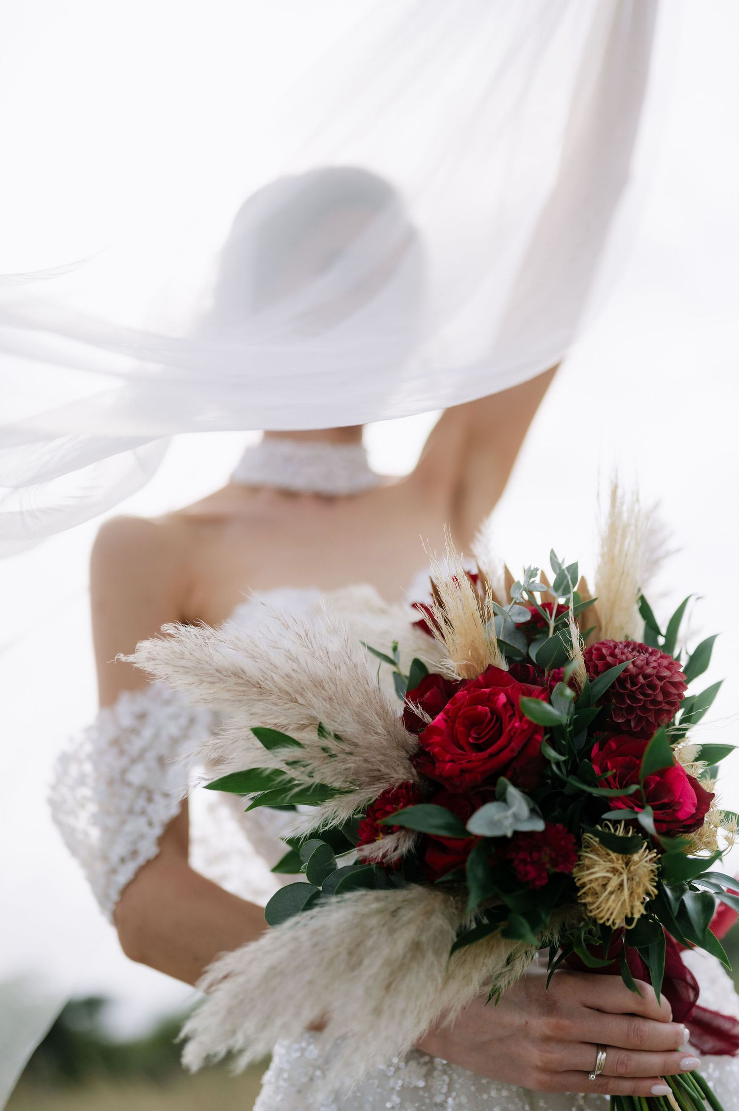 Ethereal photo of a wedding bouquet with red roses and pampas grass through flowing white veil.