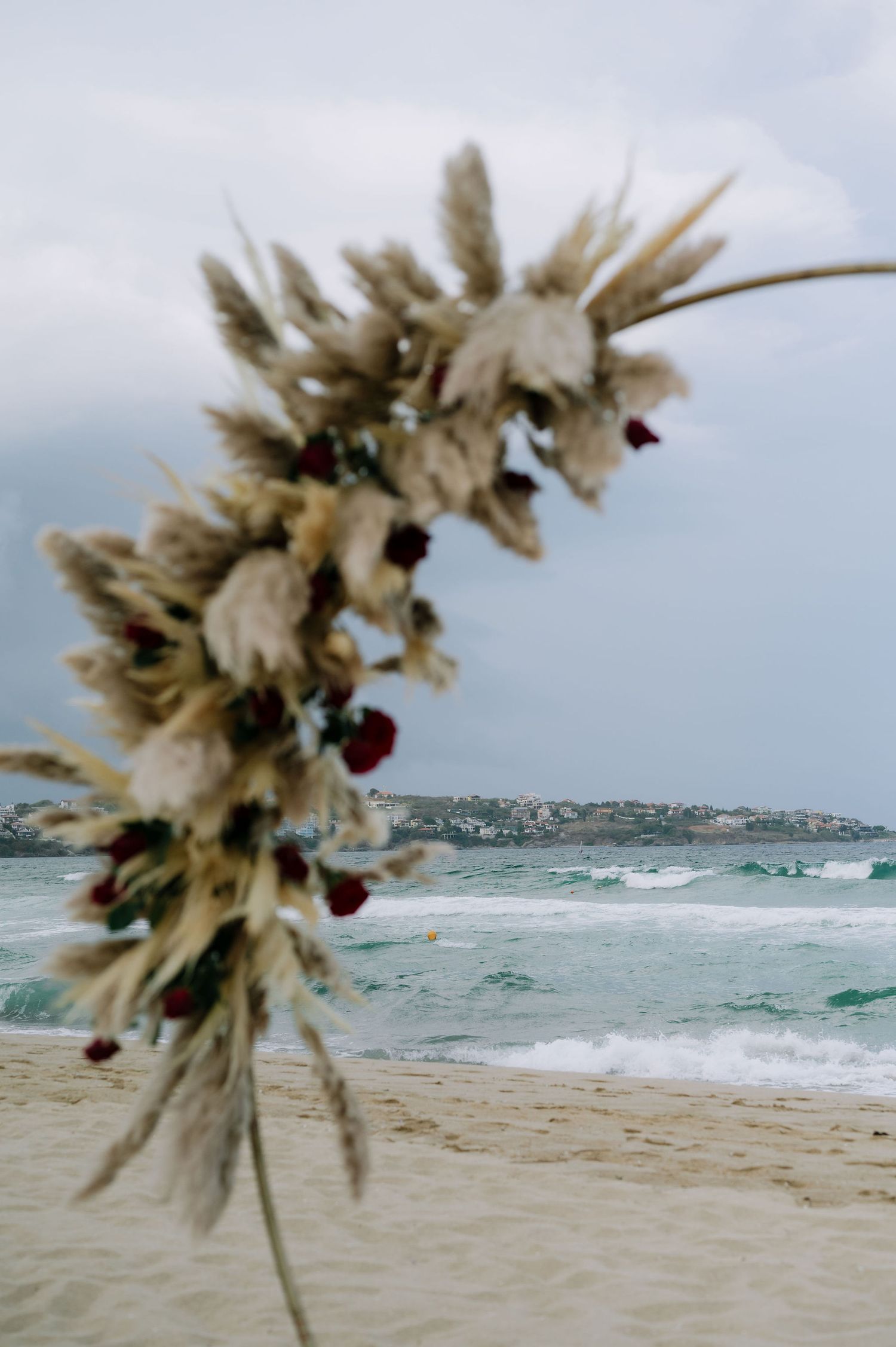 Artistic floral arrangement with pampas grass and red flowers against blurred ocean view.