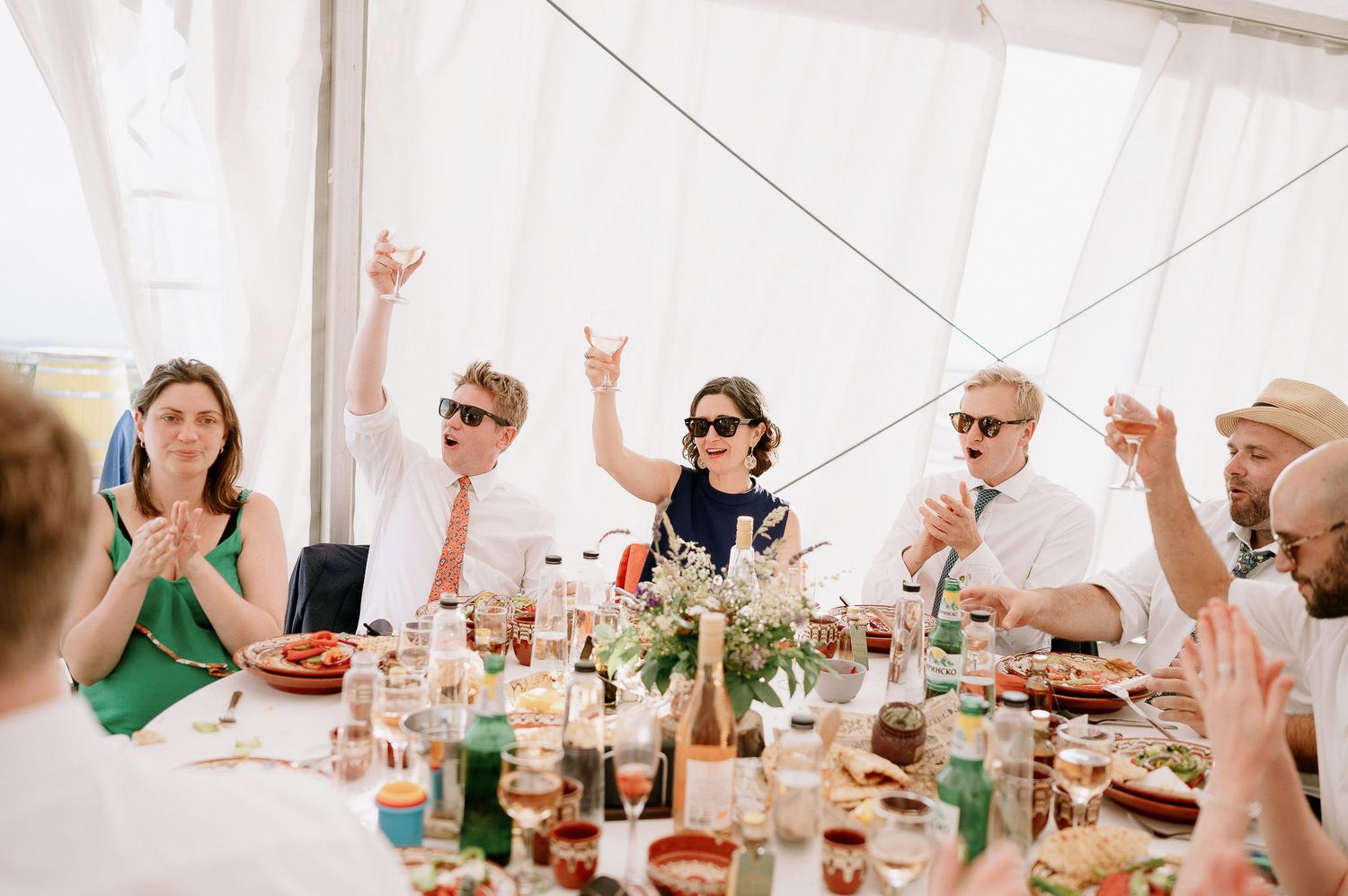 Wedding reception guests raise their glasses in celebration at a banquet table decorated with flowers and wine bottles.