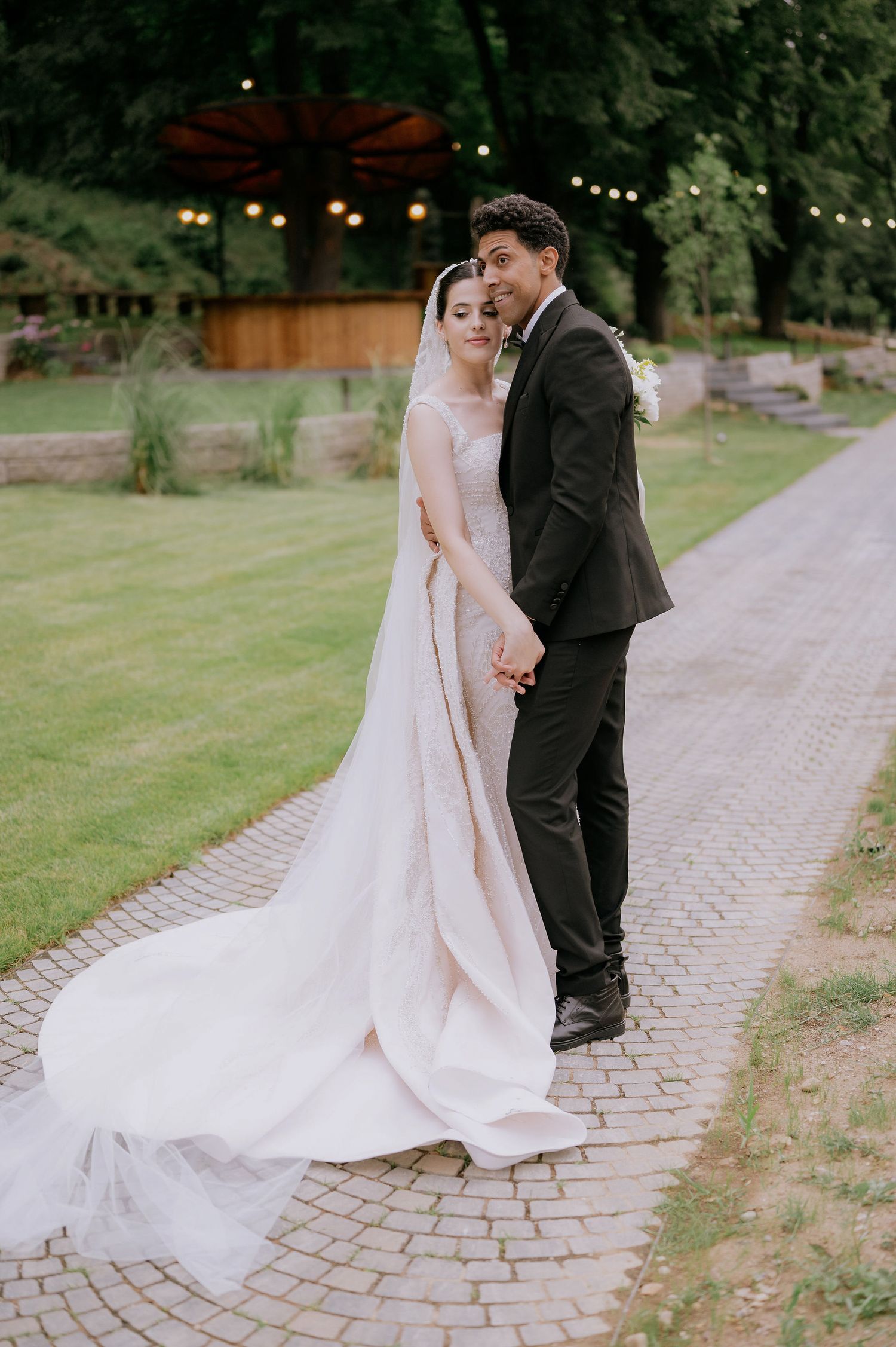 Elegant wedding couple poses on a curved brick pathway with flowing white dress and classic tuxedo.