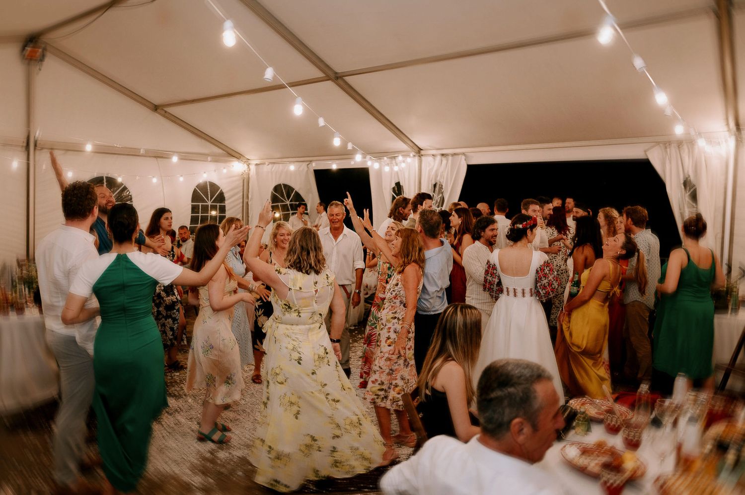 Large group of wedding guests dancing together in a white tent with string lights overhead.