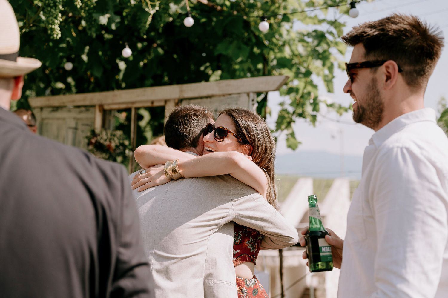 Wedding guests share an emotional embrace while holding drinks at an outdoor celebration.