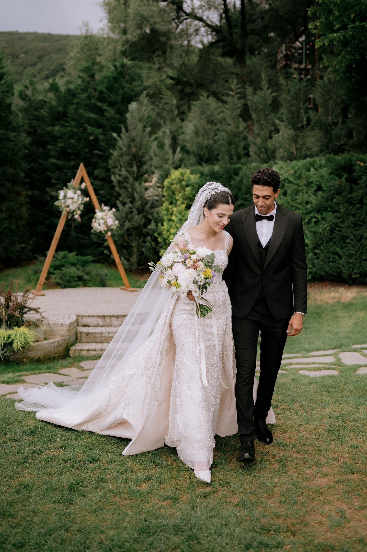 Newlyweds walking together on grass with geometric wooden arch in background.