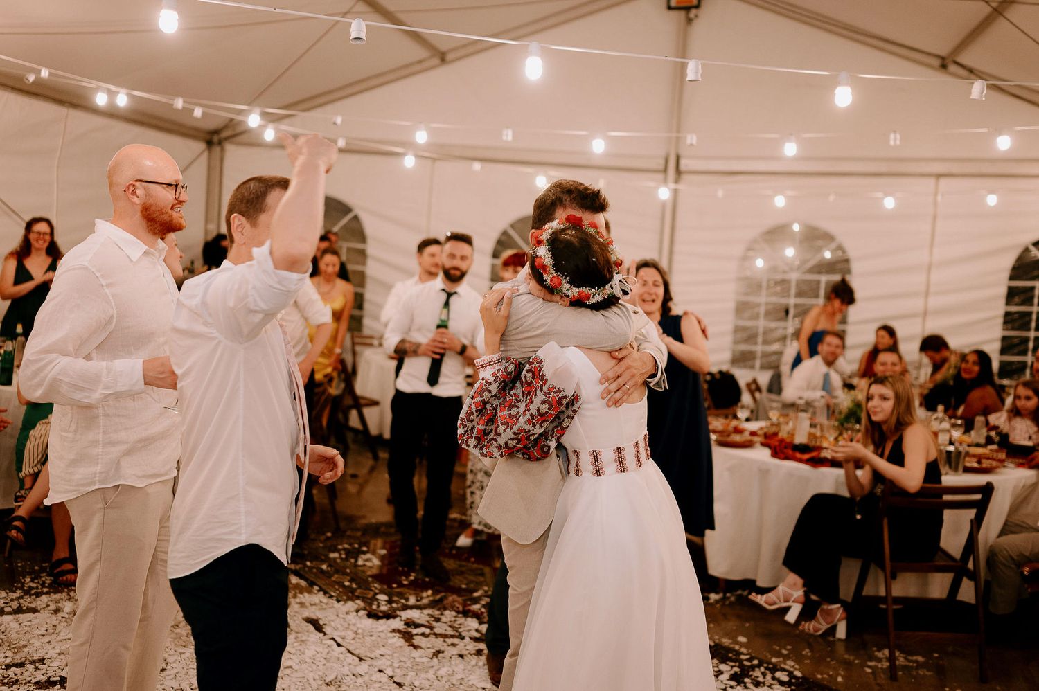 Wedding celebration with guests dancing in a tent decorated with overhead string lights.