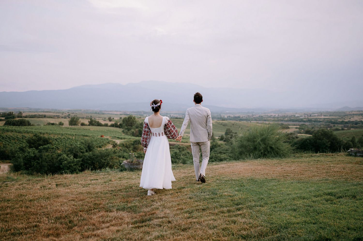 A bride in a white dress with embroidered sleeves and groom in light suit walk through a grassy meadow.