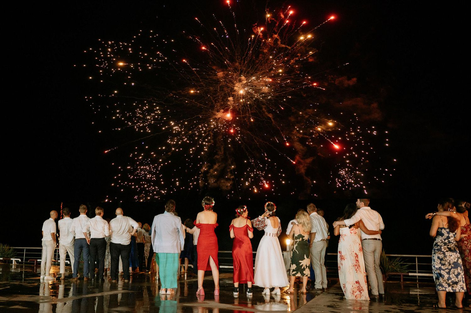 Wedding guests stand together watching a dramatic fireworks show against the dark sky.