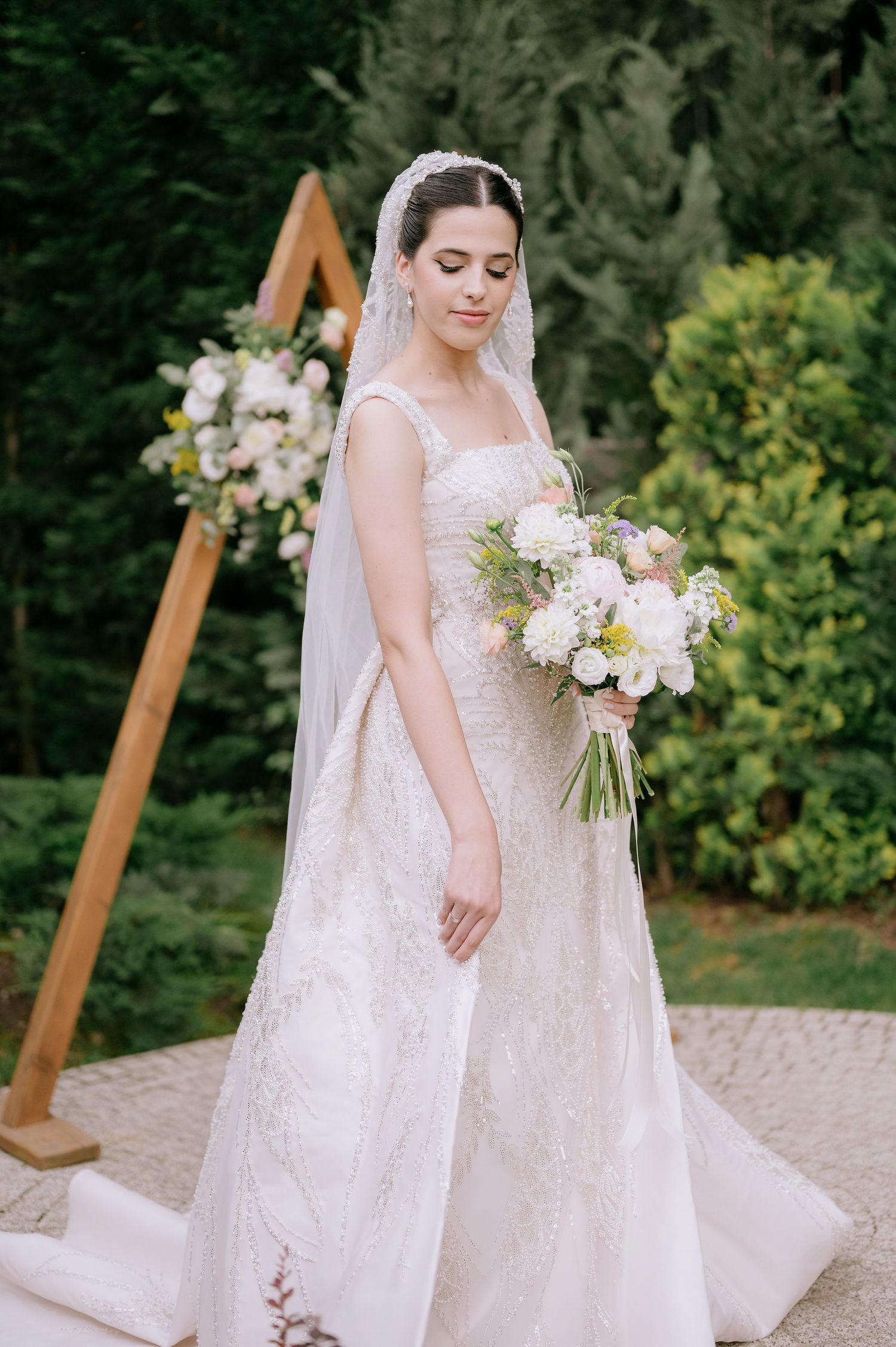 Bride in classic white gown holding pastel bouquet near wooden ceremony arch in garden setting.