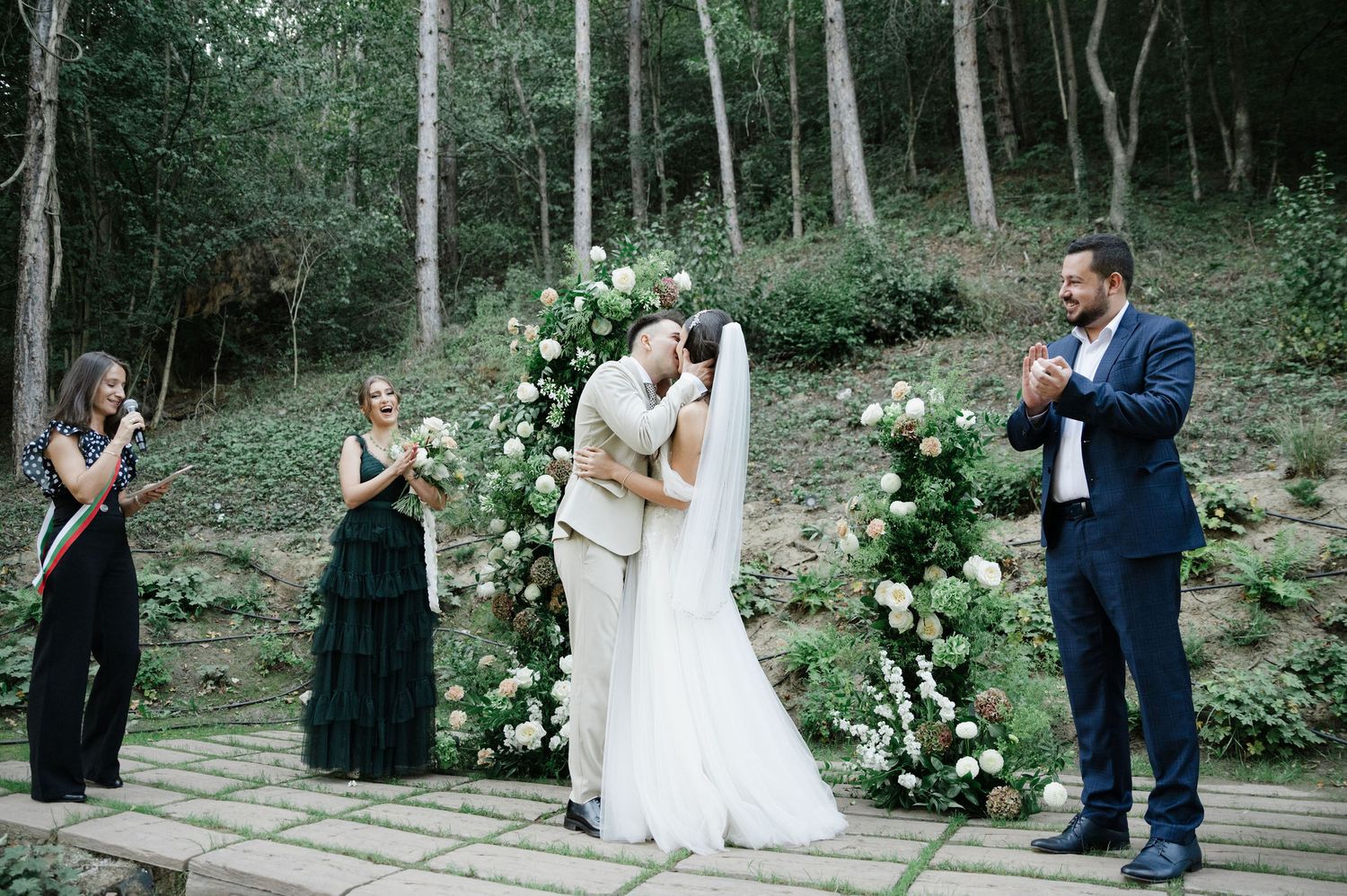Wedding ceremony under floral arch with white roses in forest setting.