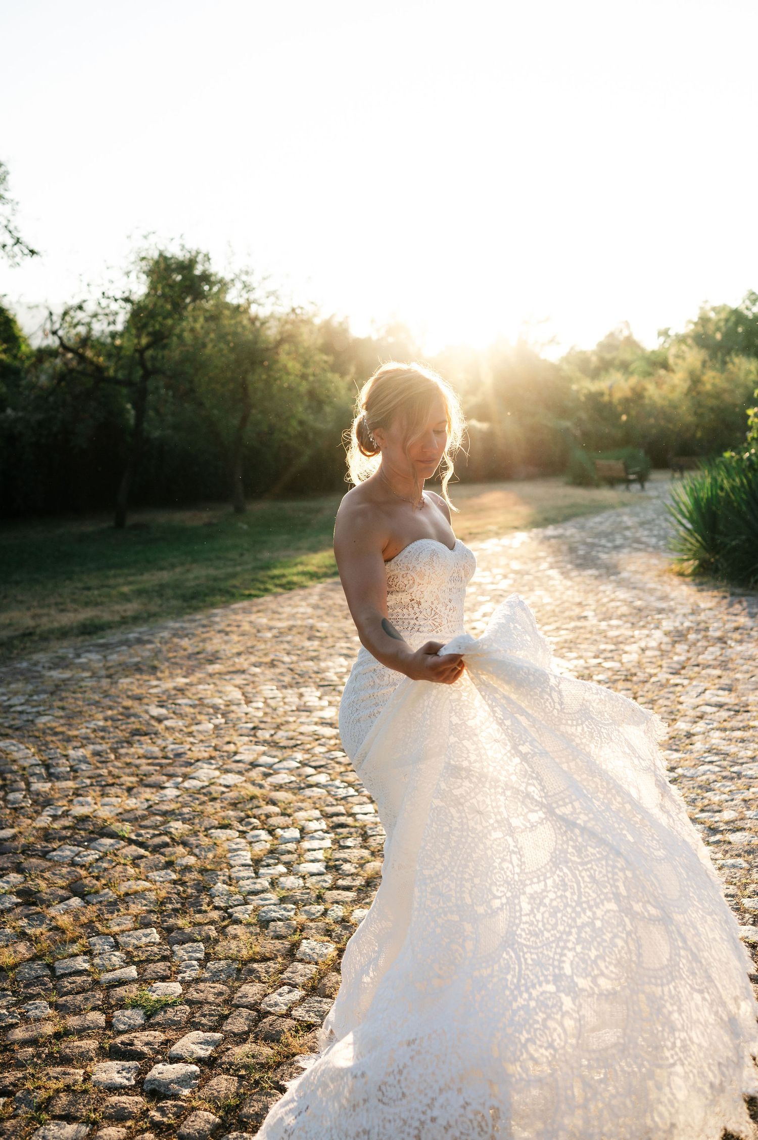 A white wedding dress flows gracefully in golden sunlight on a cobblestone path during sunset.