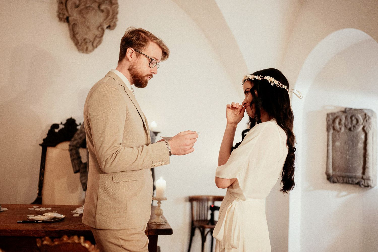 A couple in vintage-style attire share an intimate moment in a rustic white room with elegant lighting.