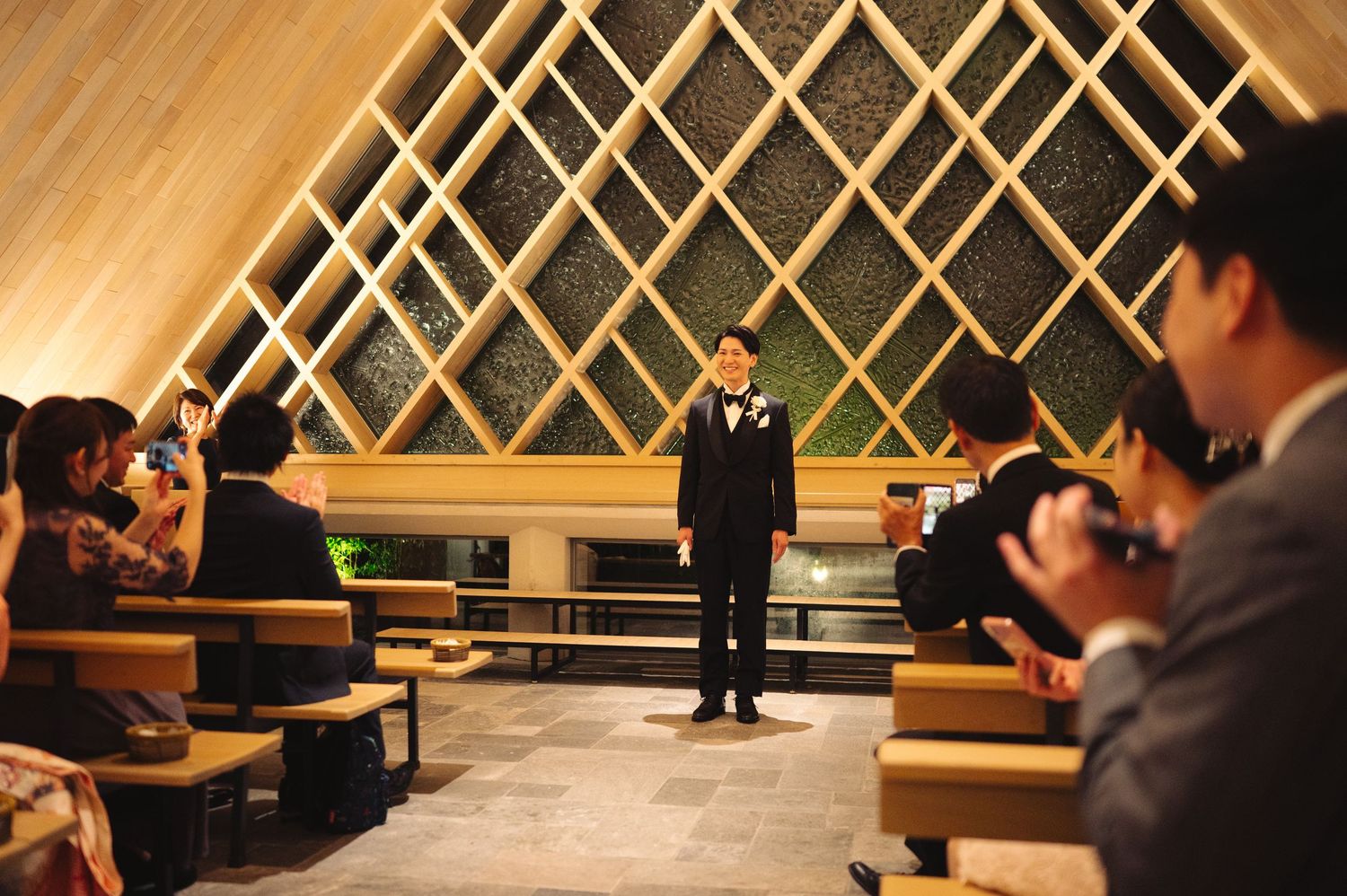 Wedding venue features geometric wooden wall panels and rows of seated attendees.