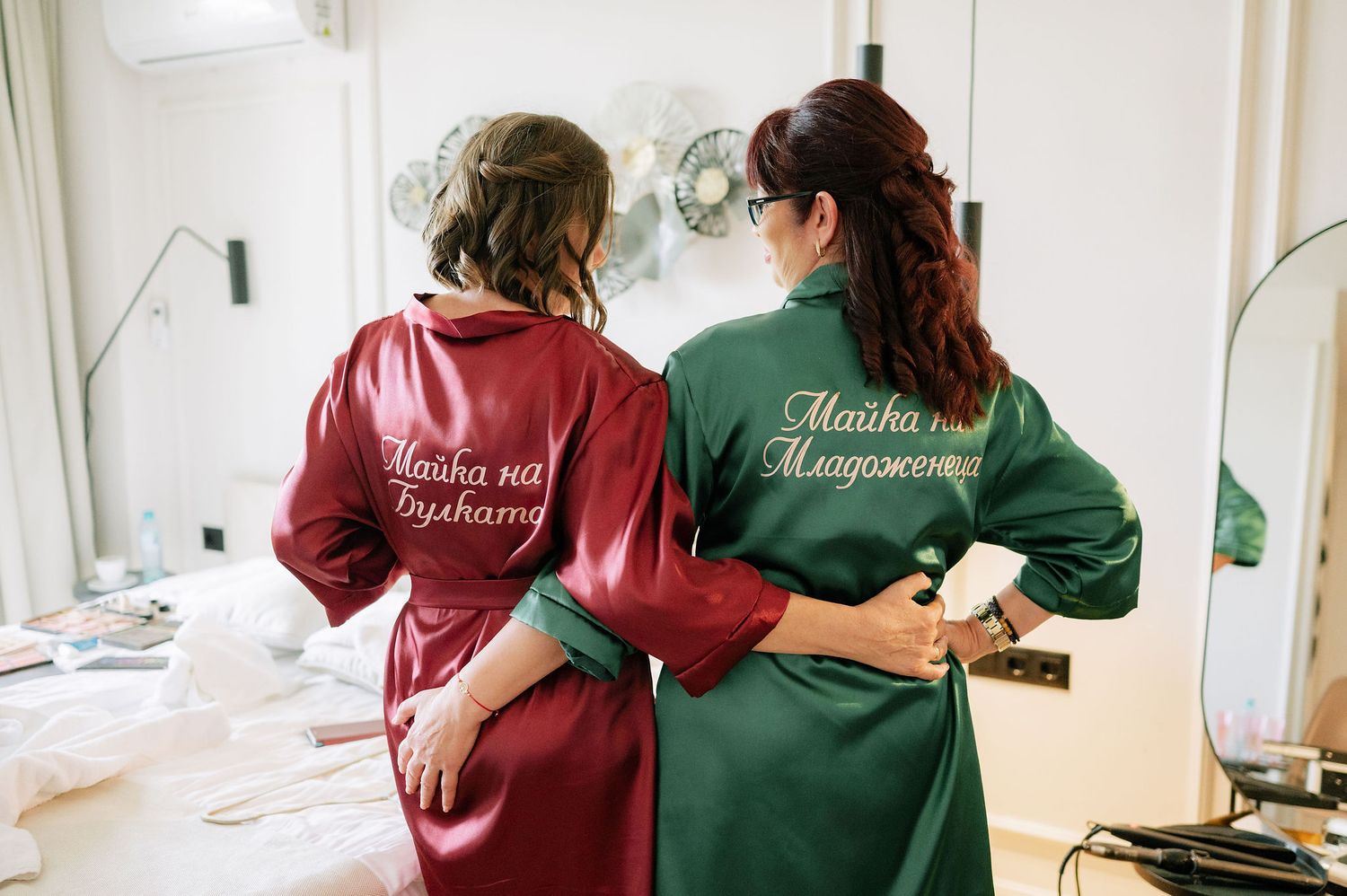 Two people in burgundy and green robes stand together during wedding preparations.