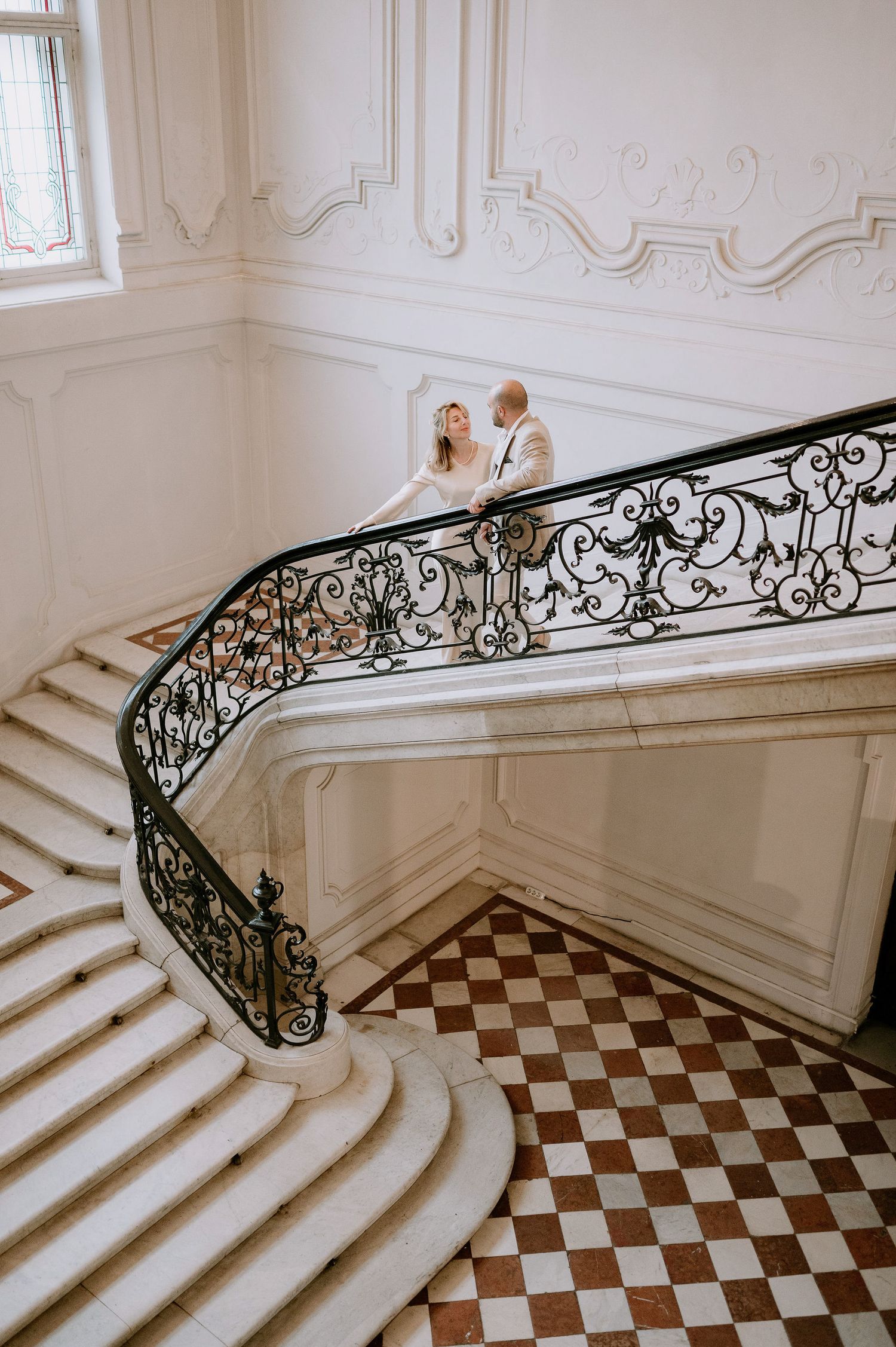 A couple stands on an elegant curved marble staircase with ornate iron railings and checkerboard floor.