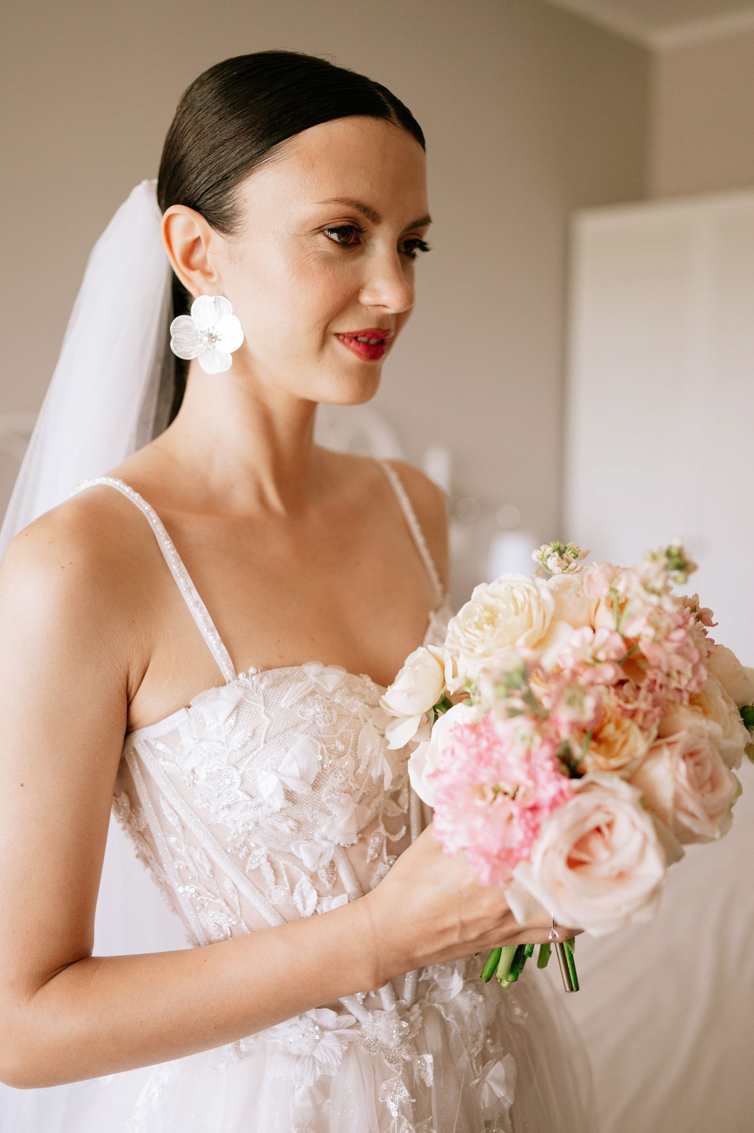 Elegant bride in lace wedding gown holds delicate pink and peach bouquet while getting ready.