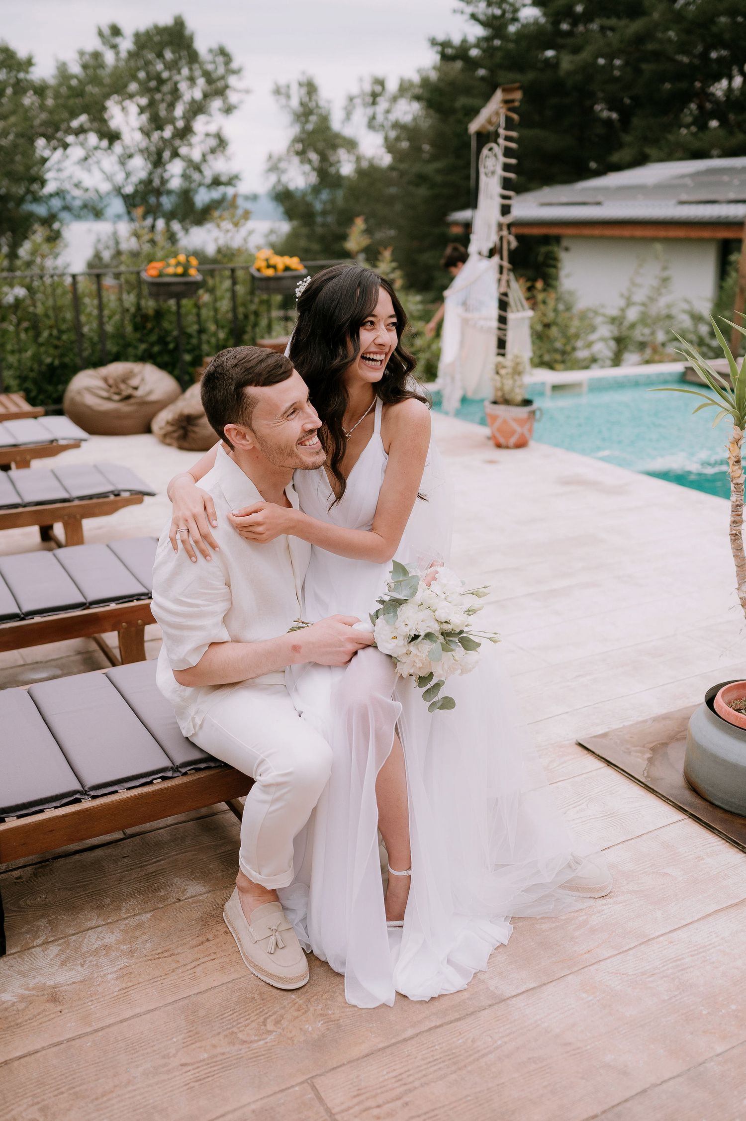 A couple in white outfits laughs together while sitting on wooden lounge chairs near a swimming pool.