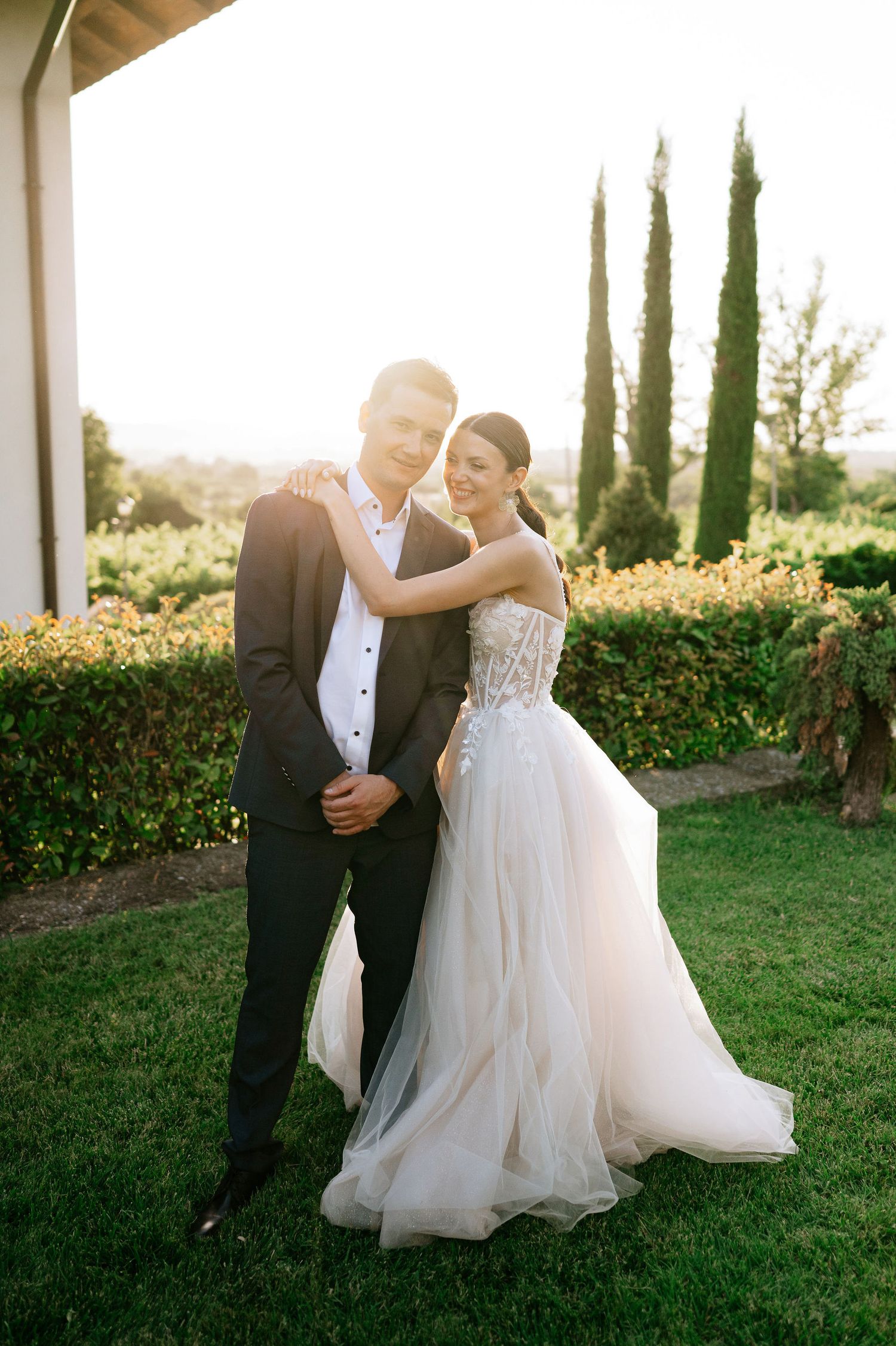 Wedding couple embraces while dancing on a manicured lawn during sunset with Italian cypress trees.