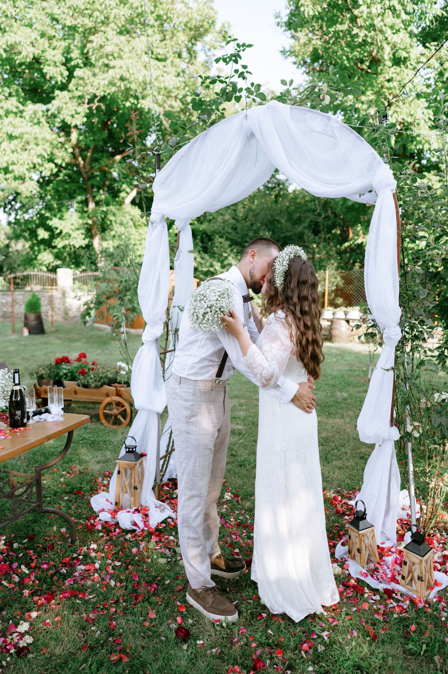 An intimate wedding moment under a white fabric arch with scattered rose petals on the ground.