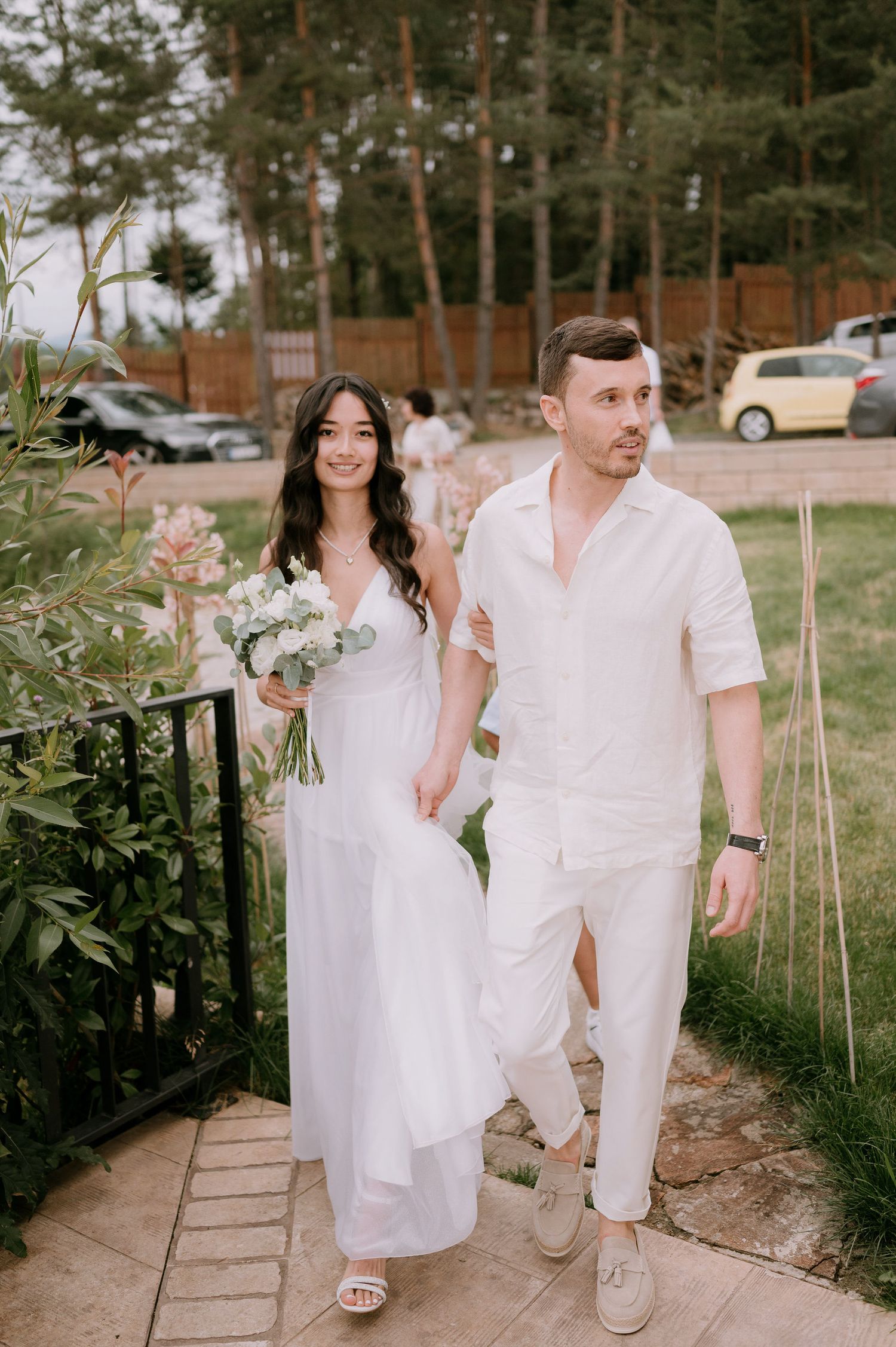 Wedding couple strolling along garden path in coordinated white outfits with floral bouquet.