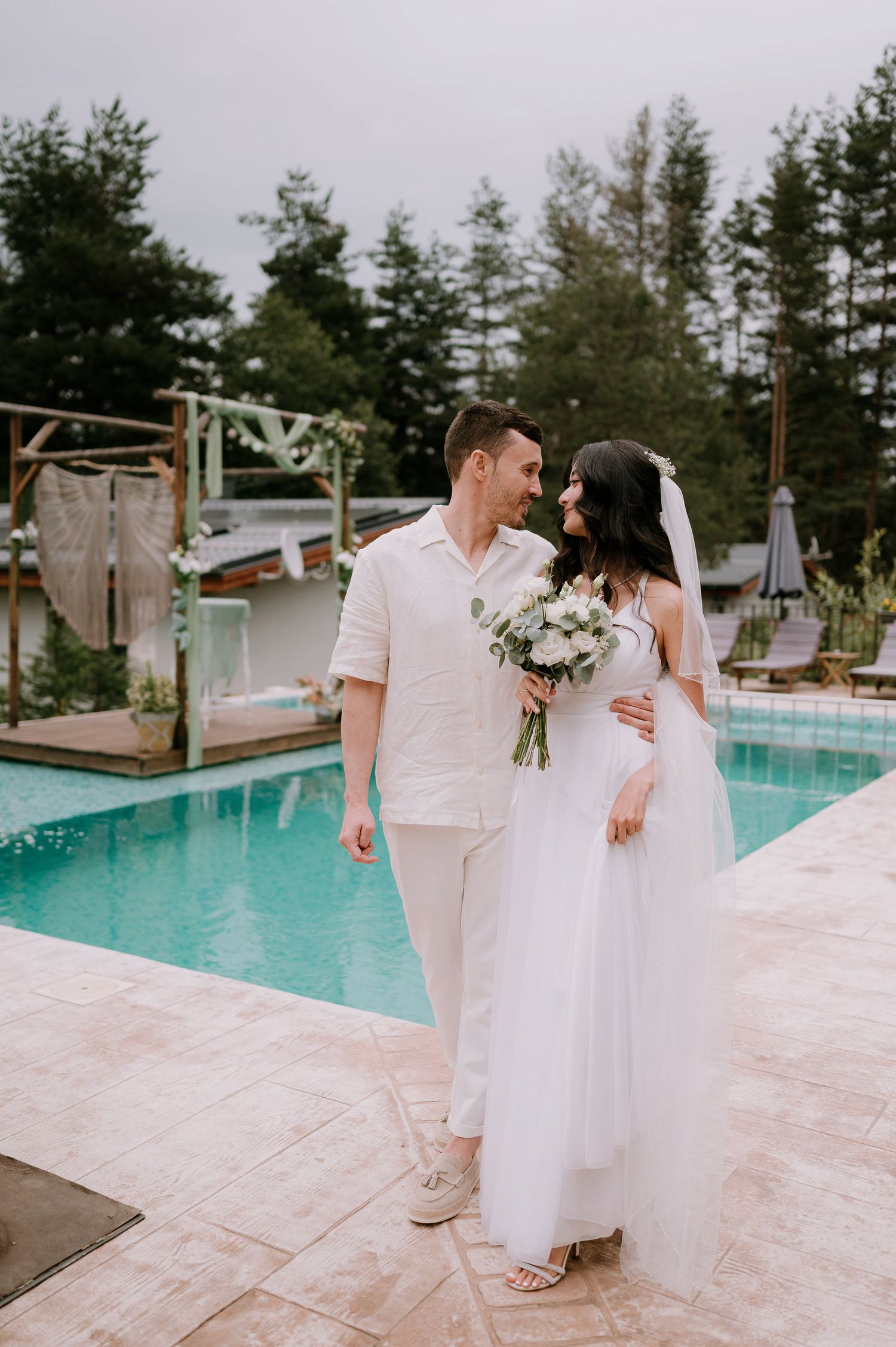 A romantic wedding celebration unfolds beside a pristine pool with pine trees in the background.