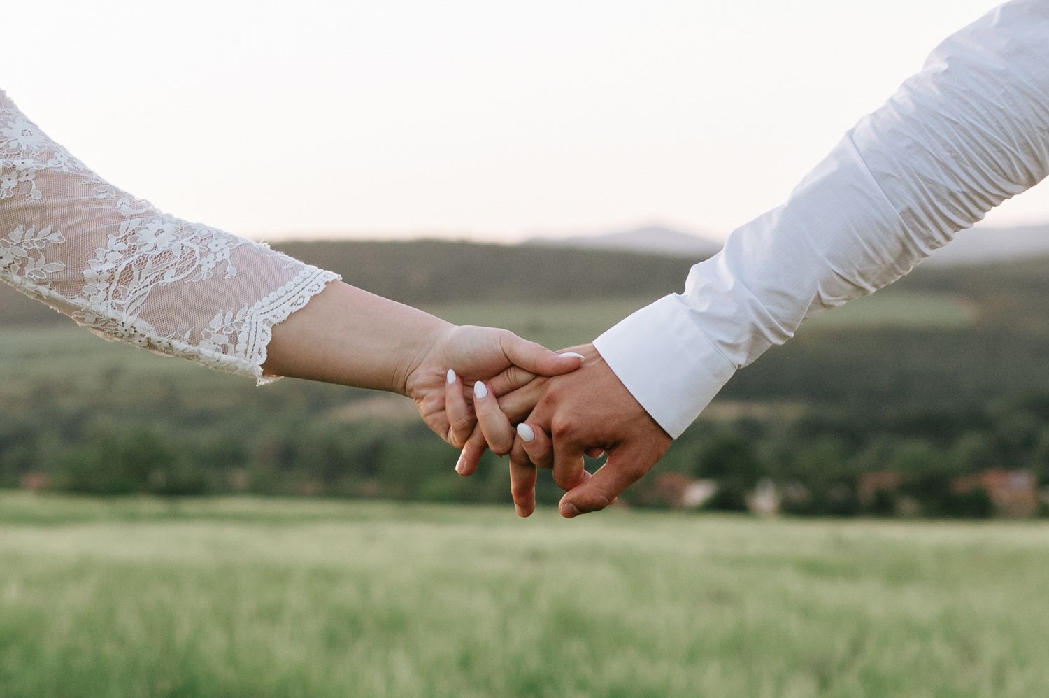 Close-up of hands reaching for each other against a sunset backdrop in a grassy field.