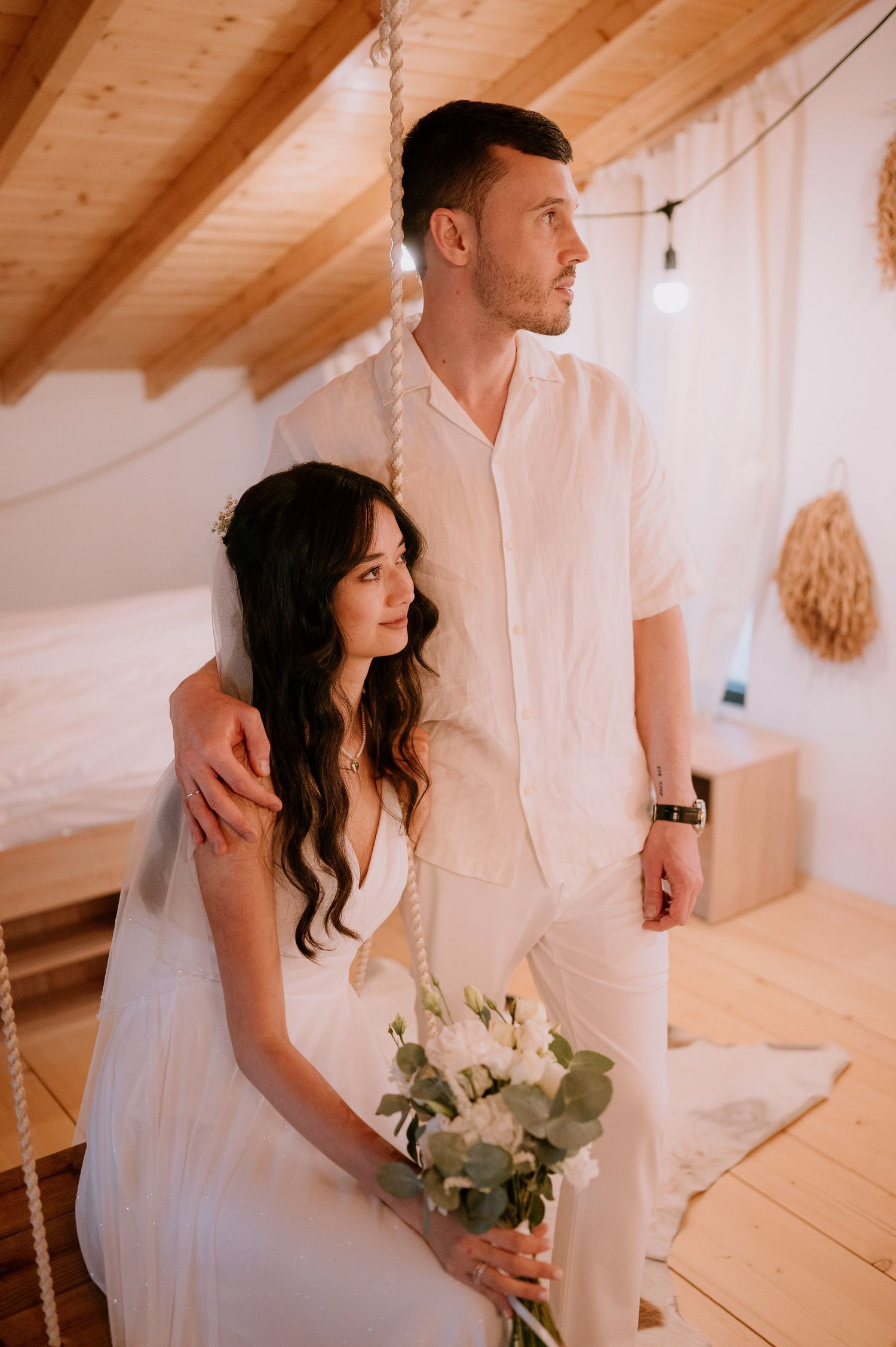 Couple in all-white attire posing in rustic indoor setting with wooden ceiling beams.