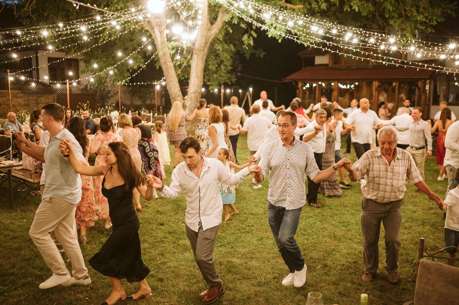 Joyful wedding celebration with guests dancing on grass under glowing string lights.