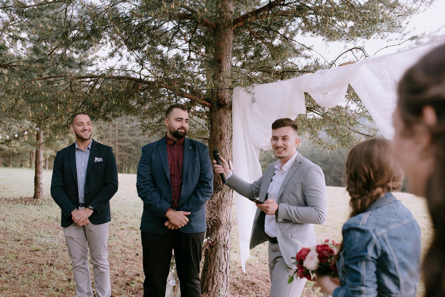 Wedding party members gather under pine trees for a casual outdoor ceremony.