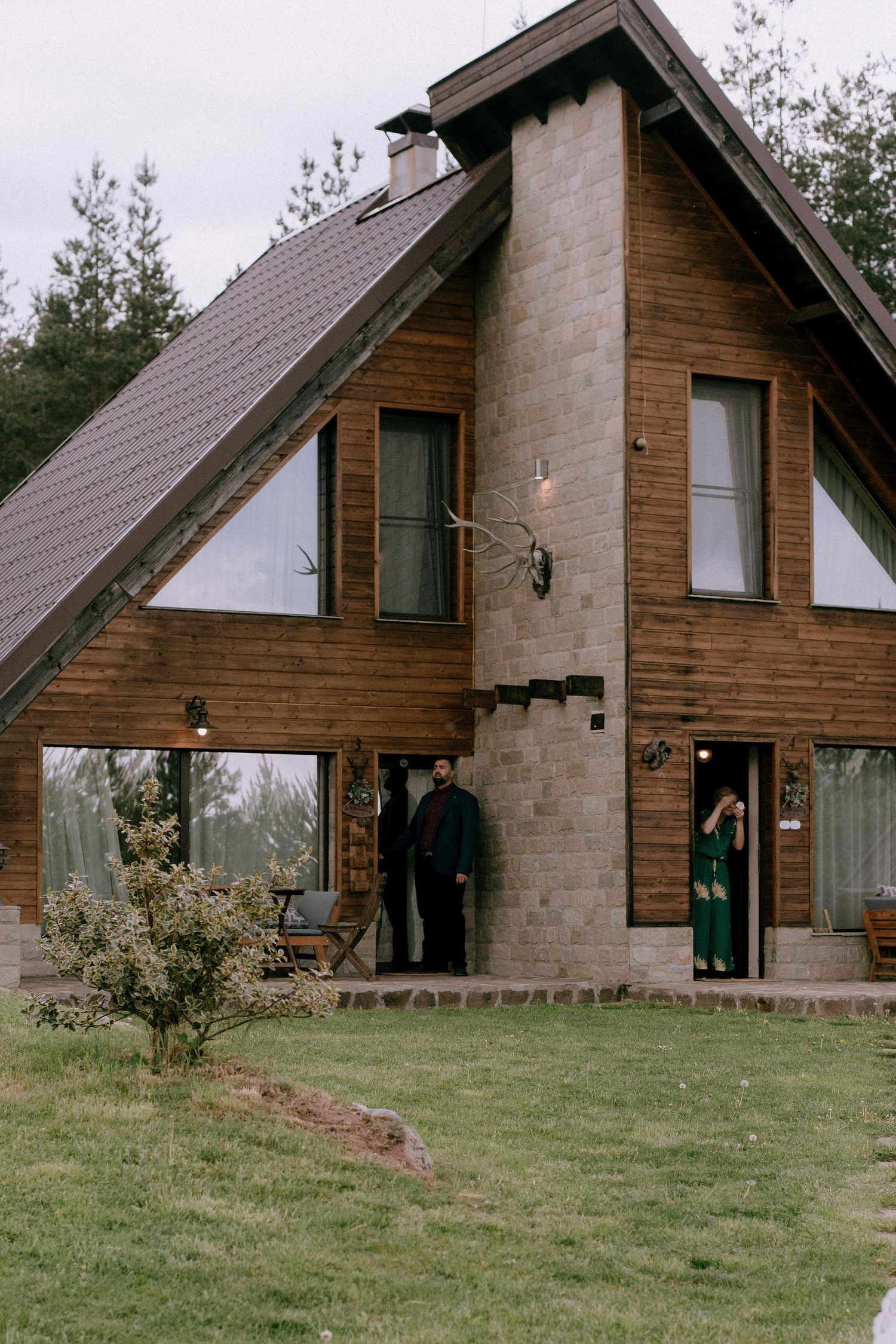 A rustic A-frame cabin with wood and stone exterior sits on a grassy hillside.