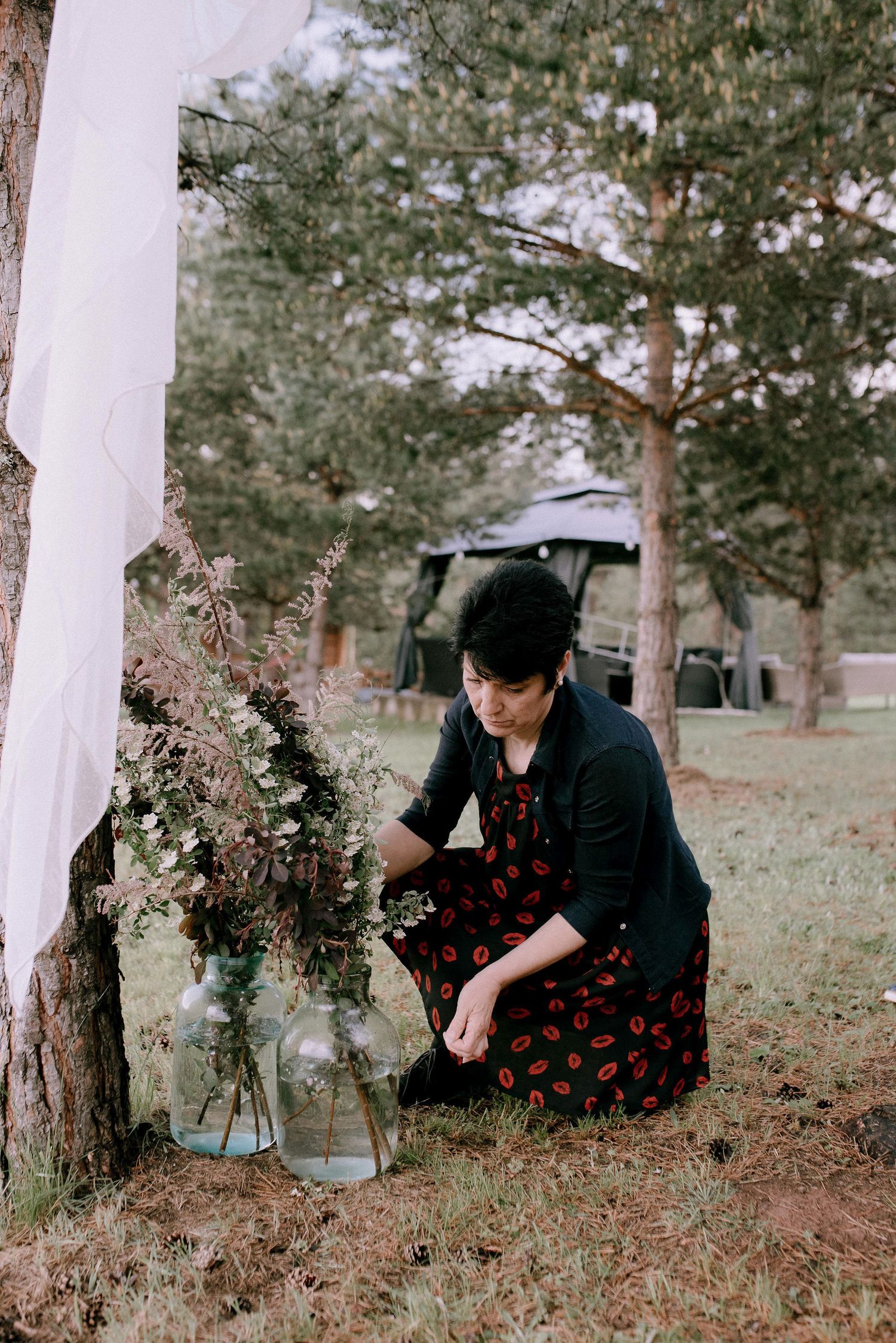 Person in dark clothing arranging flowers in glass vases outdoors near pine trees.