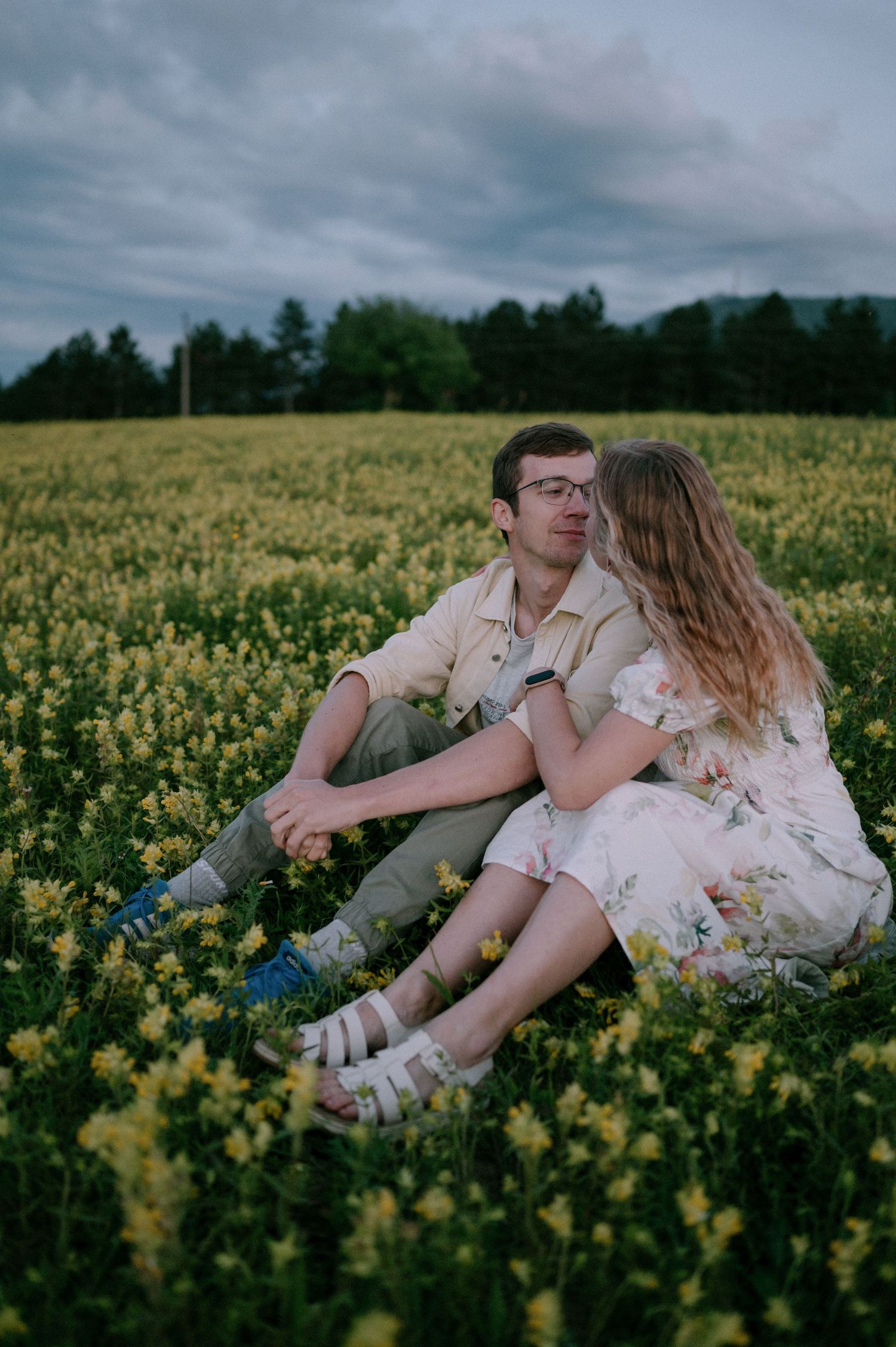 A couple shares an intimate moment while lying in a field of yellow flowers under cloudy skies.