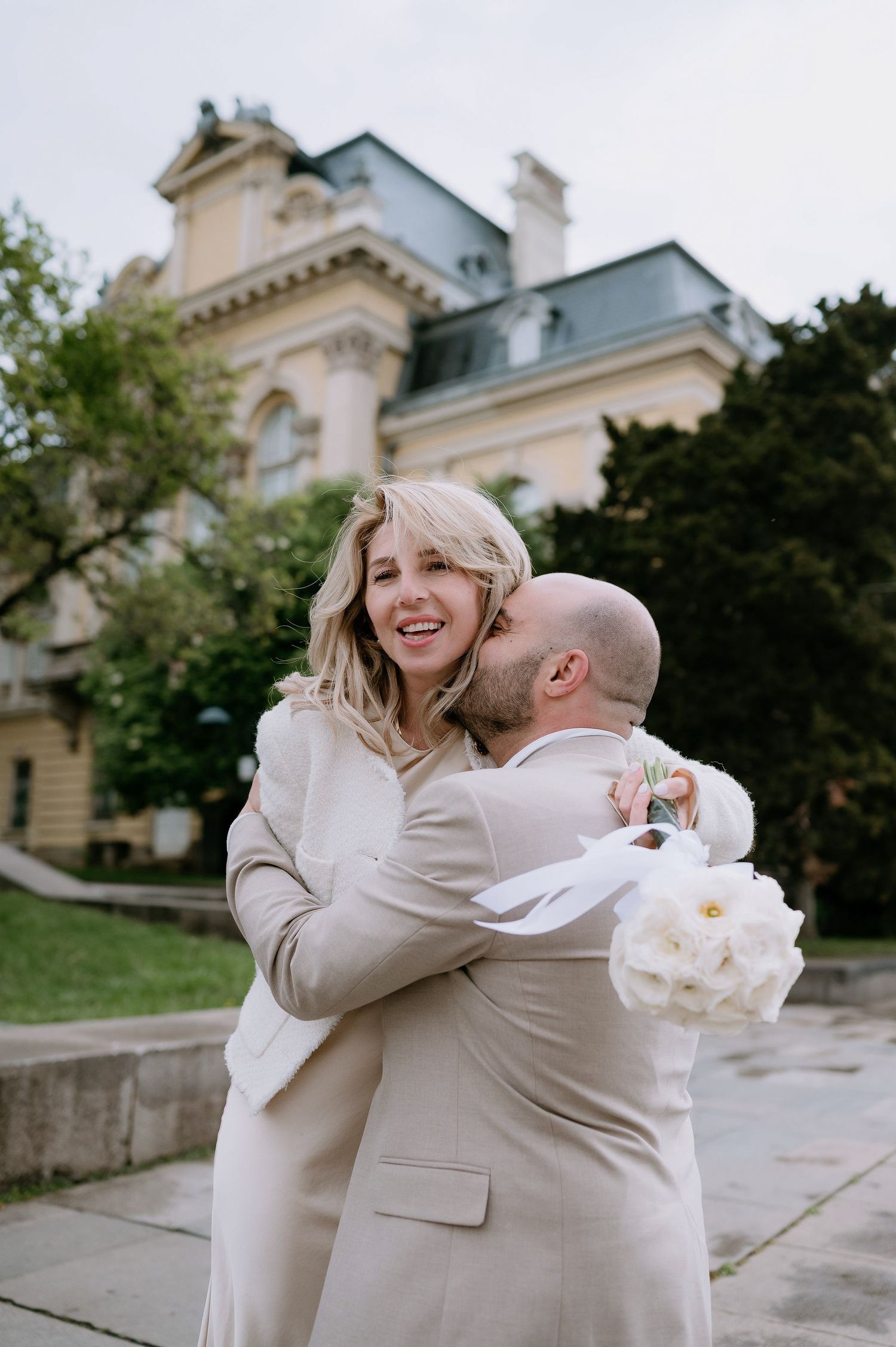 A couple dressed in cream-colored outfits embrace lovingly outside an elegant building.
