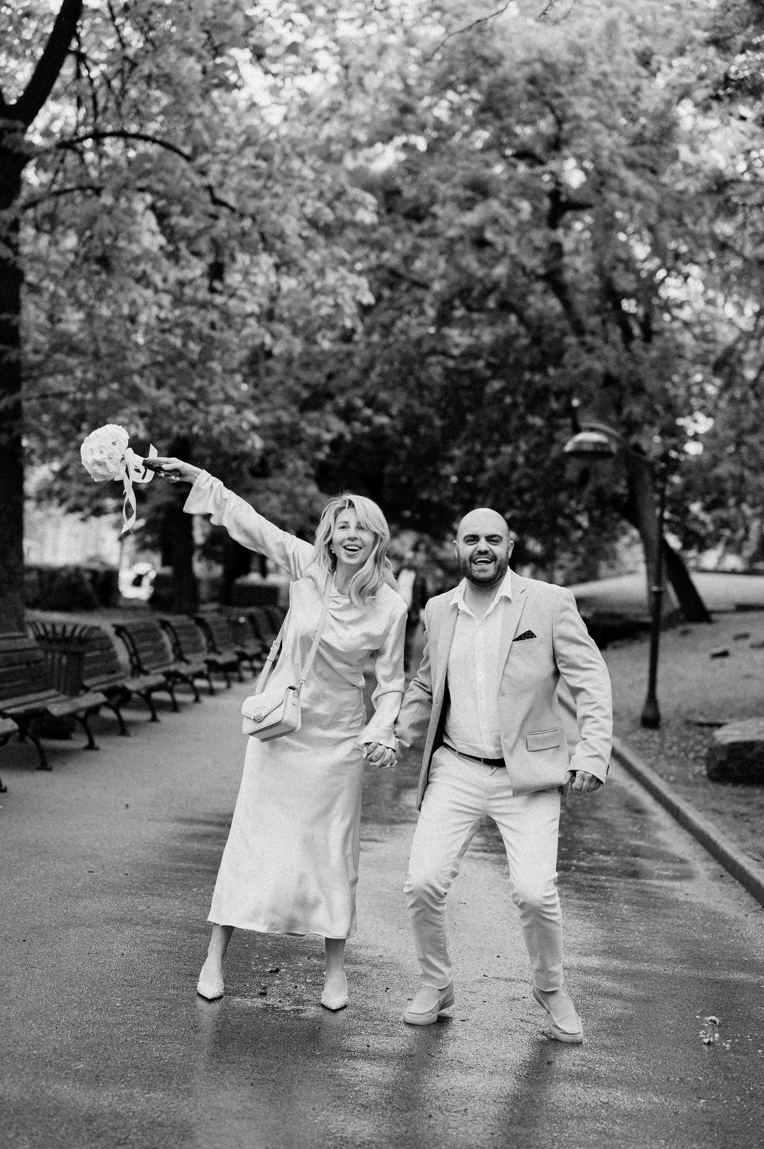 A joyful couple in light-colored clothing shares a dance moment on a rainy park path.