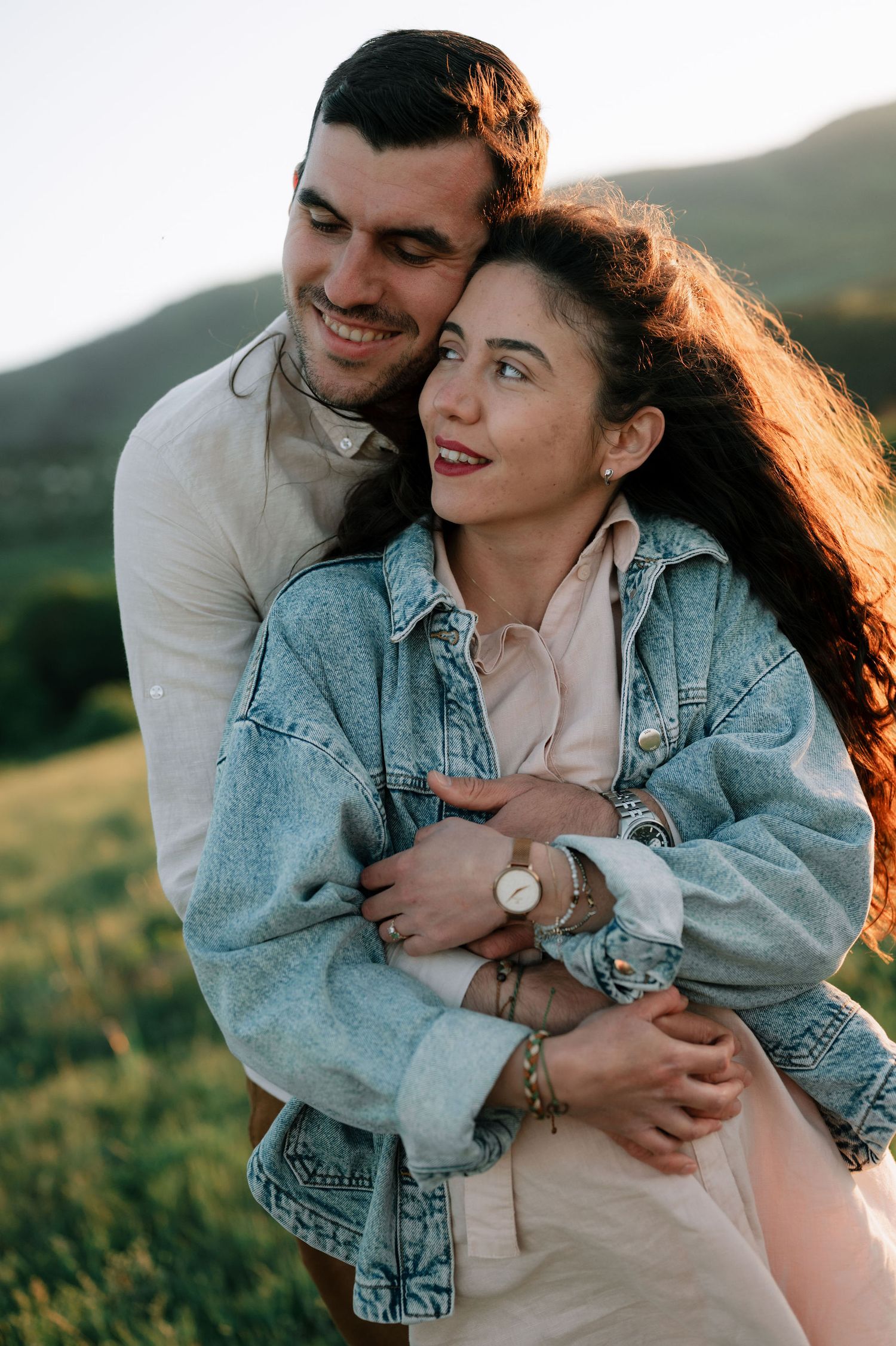 A romantic couple embraces on a hillside at sunset, with her wearing a denim jacket and him in white.
