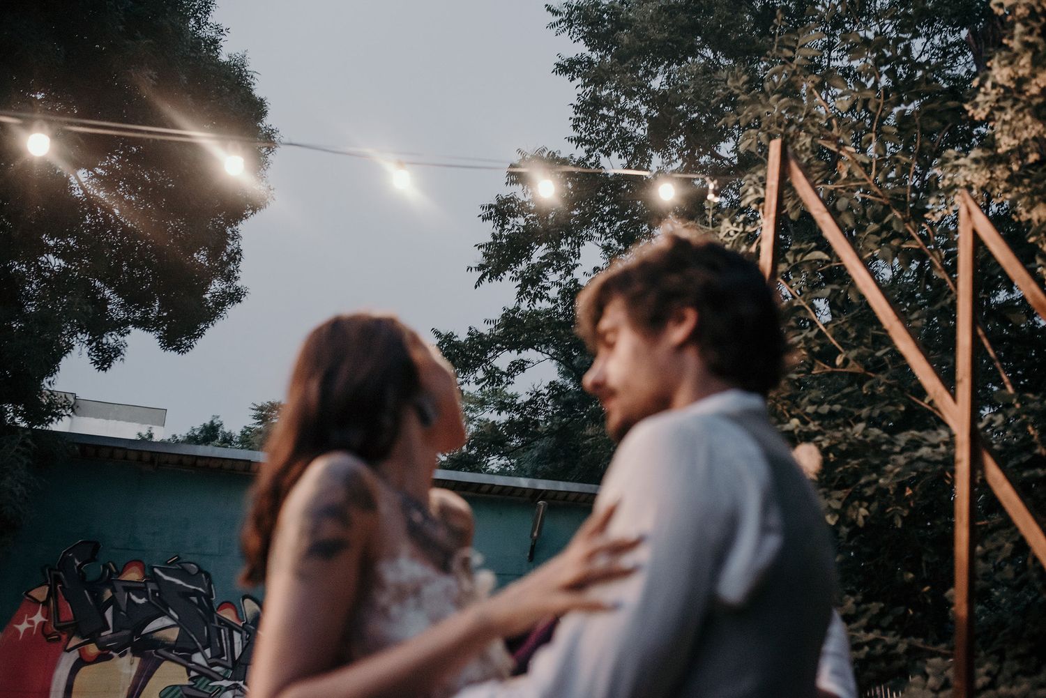 A couple embraces under a rustic wooden structure at dusk with trees in the background.