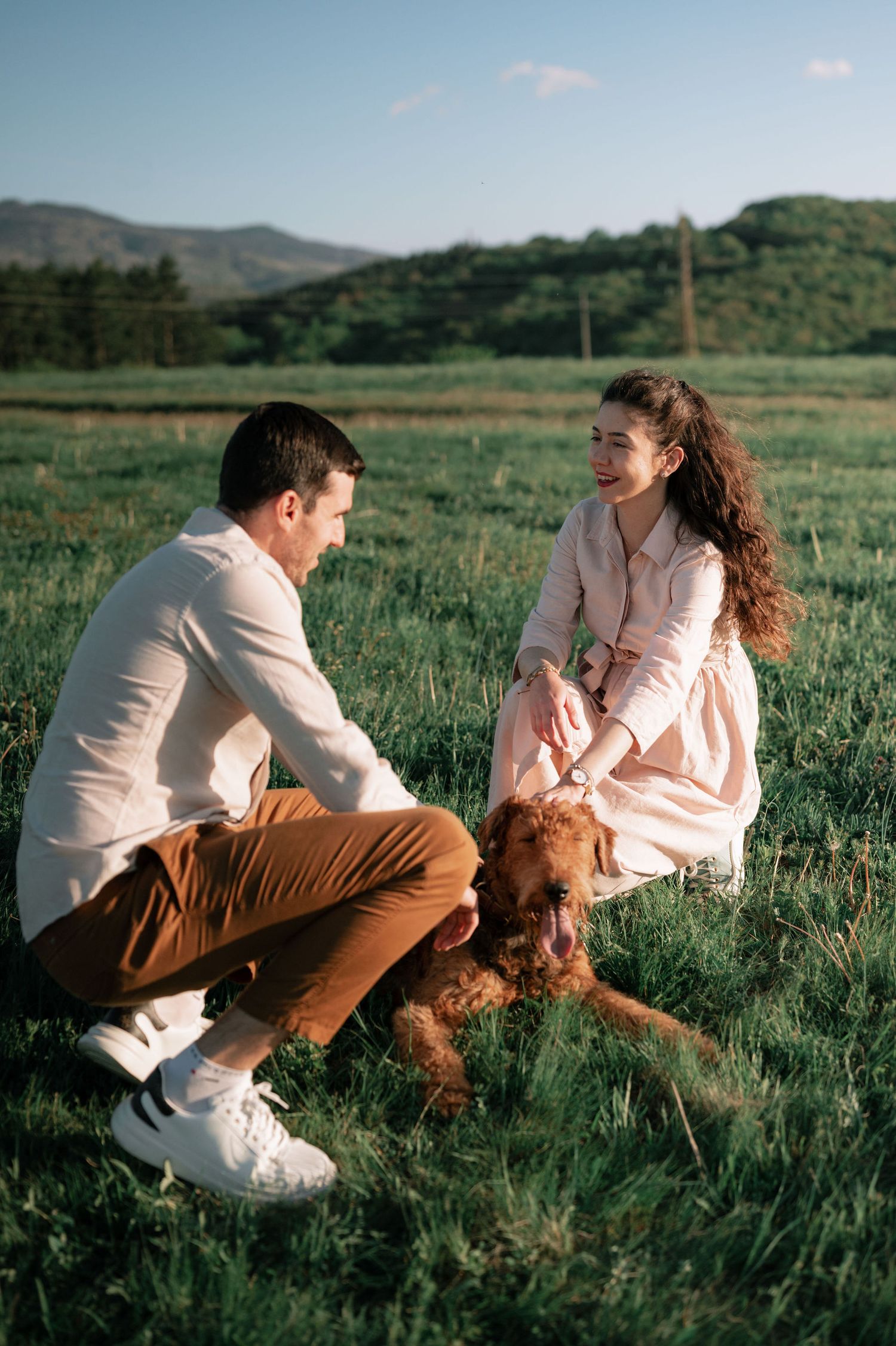 A couple sits in a grassy meadow with their golden retriever dog as mountains rise in the background.