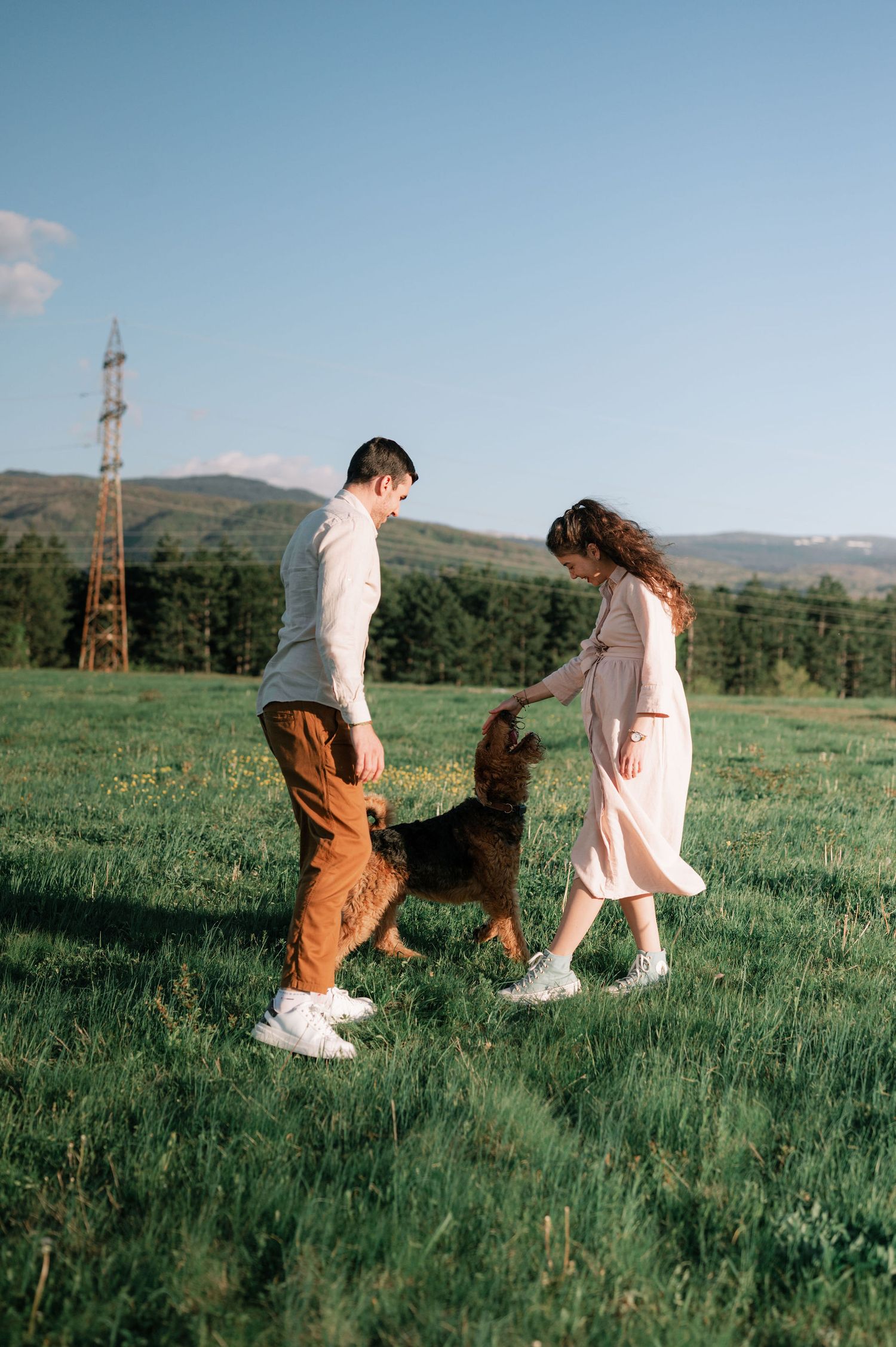 Two people in coordinating outfits walk together through a lush green field with mountains and transmission towers in background.