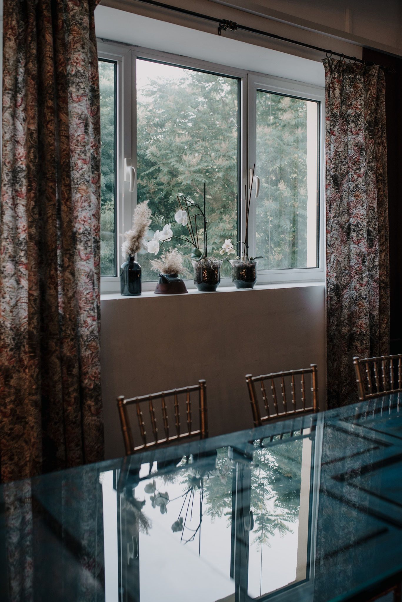 Elegant dining room window with floral curtains and potted plants on the sill reflecting in a glass table.