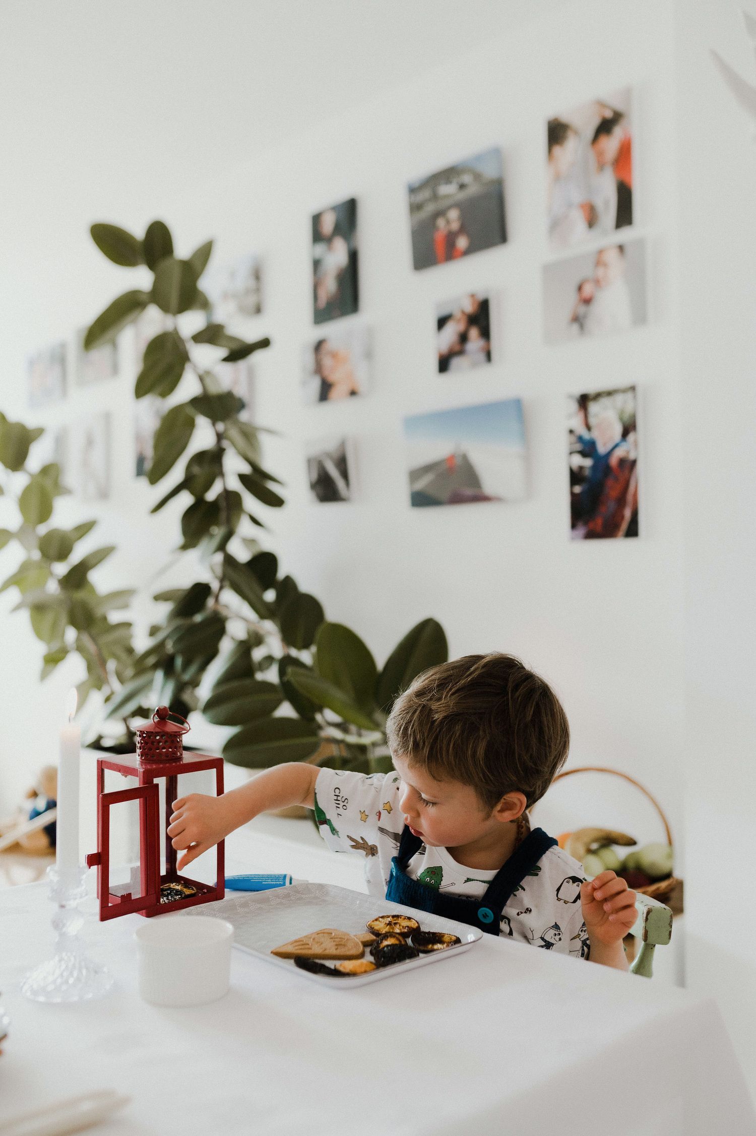 A child sits at a white table with photos displayed on the wall and greenery in view.