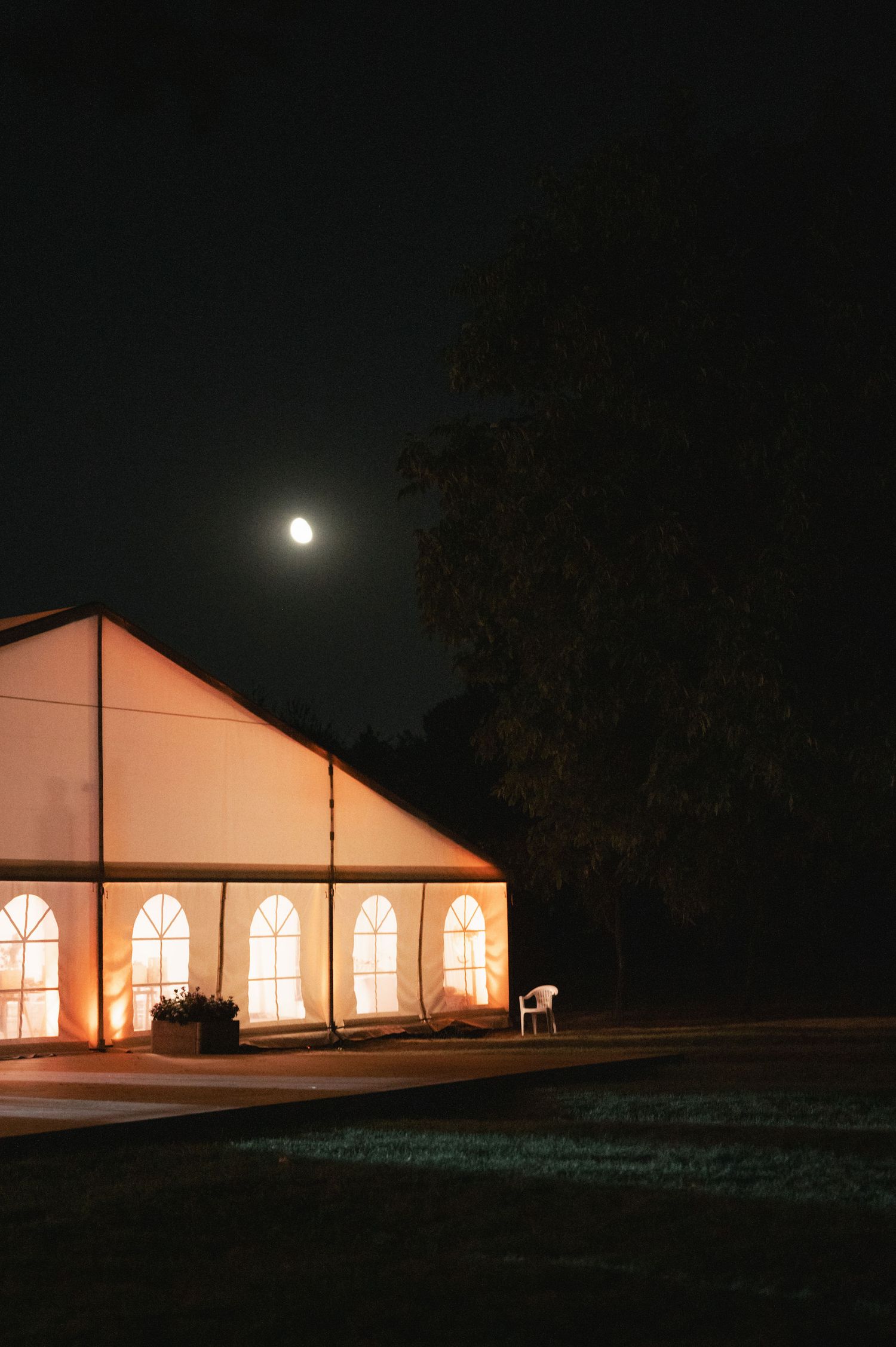Illuminated white wedding tent glows warmly against dark night sky with full moon visible above trees.