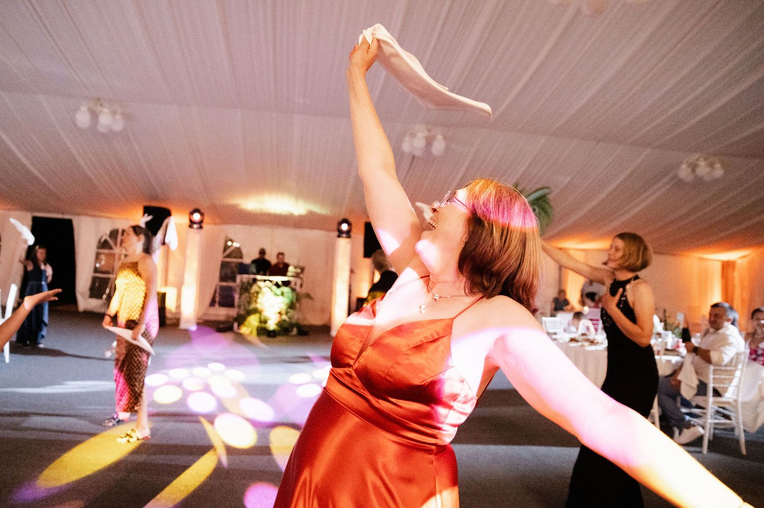 People dancing with raised arms on a dance floor at a wedding reception with white tent ceiling and lighting.