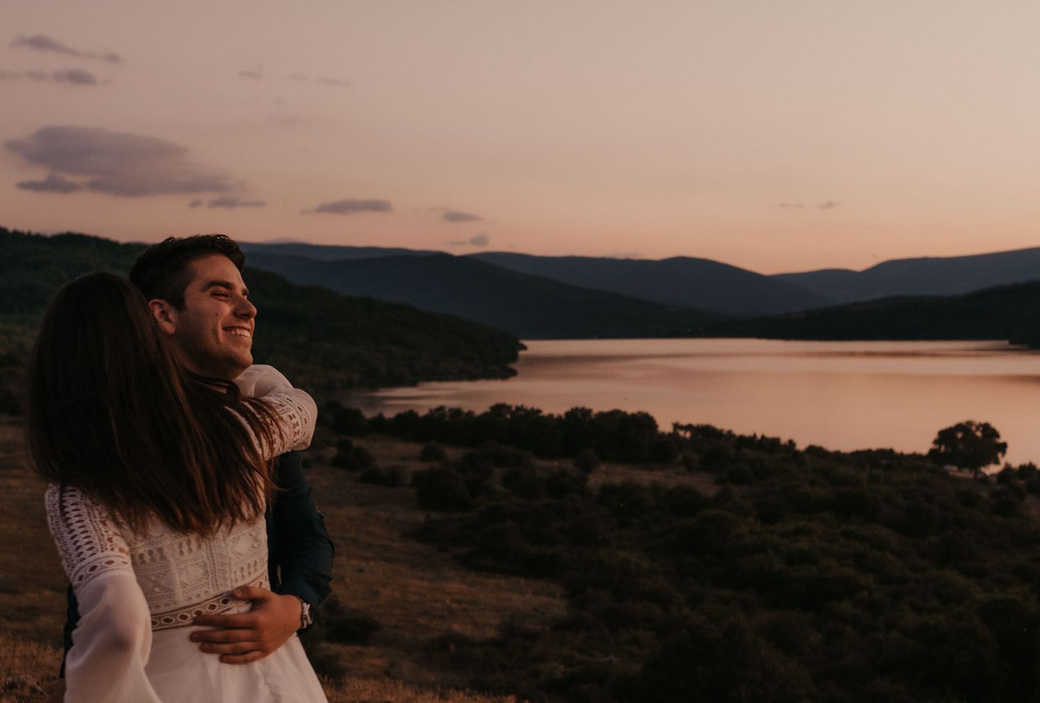 A couple stands together on a grassy hillside during golden hour with mountains and water in the distance.