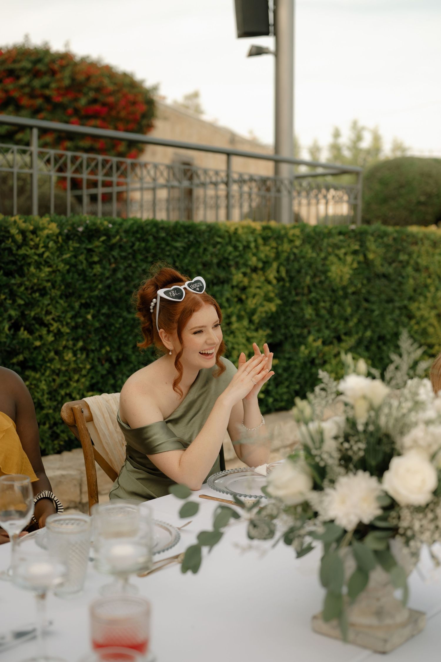 Red-haired woman in olive off-shoulder dress with sunglasses on head, smiling at outdoor wedding reception table with white flowers.