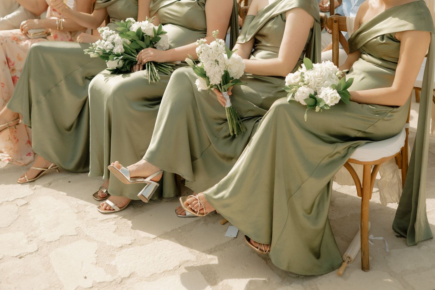 Bridesmaids in sage green satin dresses holding white floral bouquets seated in chairs at an outdoor wedding ceremony.