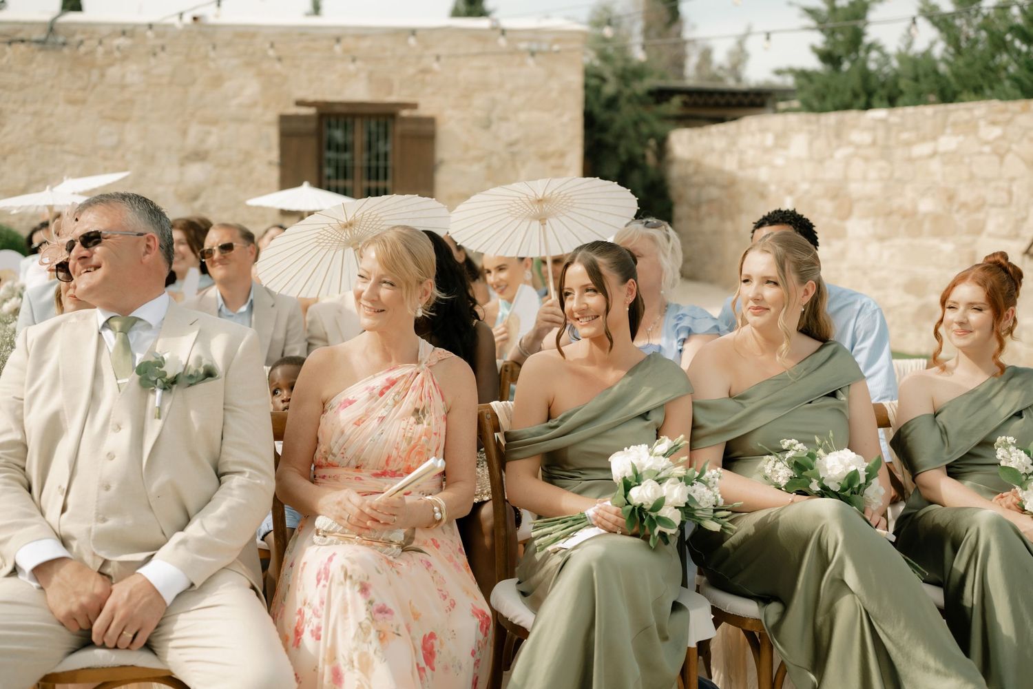 Wedding guests in sage green bridesmaid dresses holding white bouquets seated outdoors at an elegant summer ceremony.