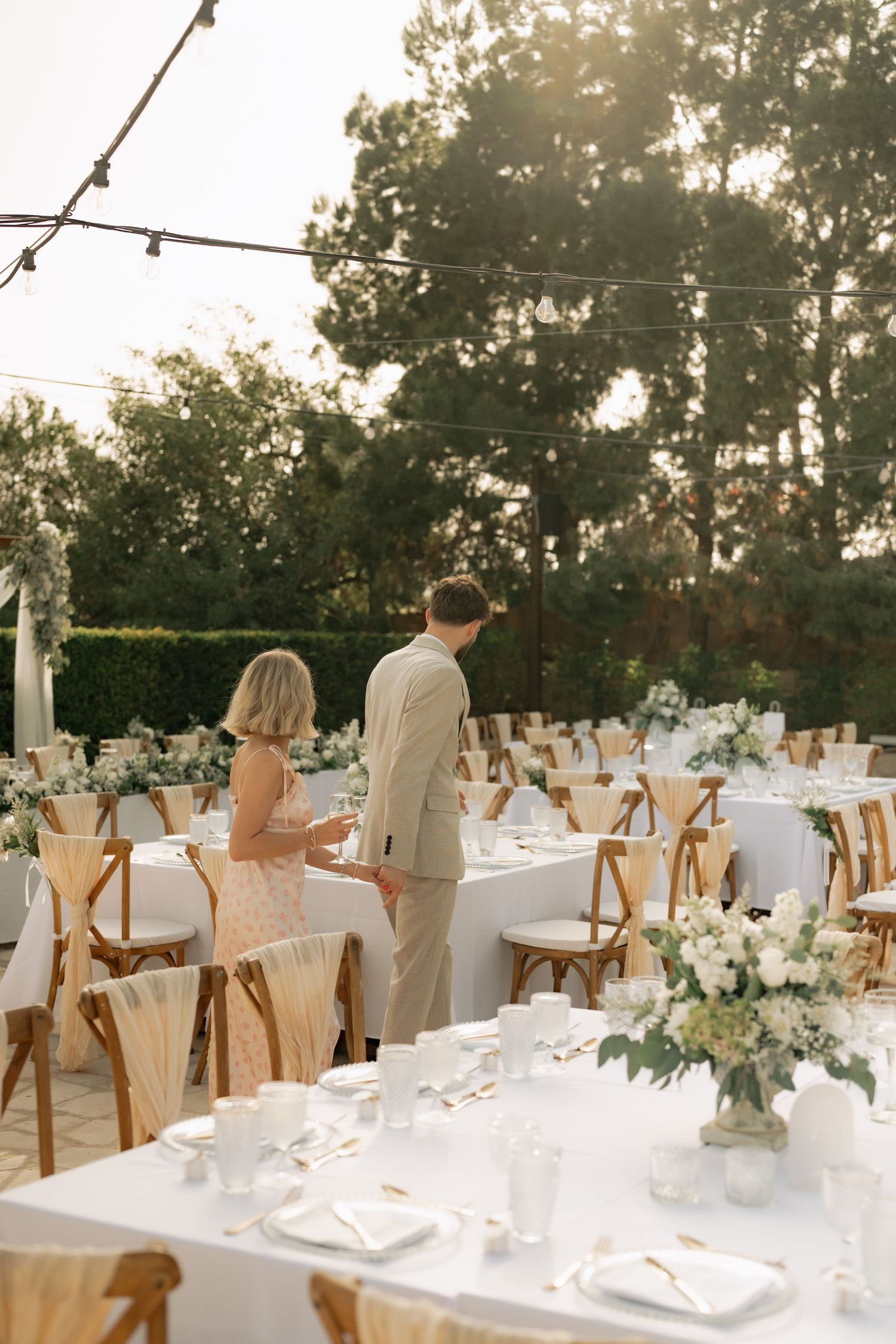 Elegant outdoor wedding reception with white tablecloths, floral centerpieces, and couple standing among wooden cross-back chairs.