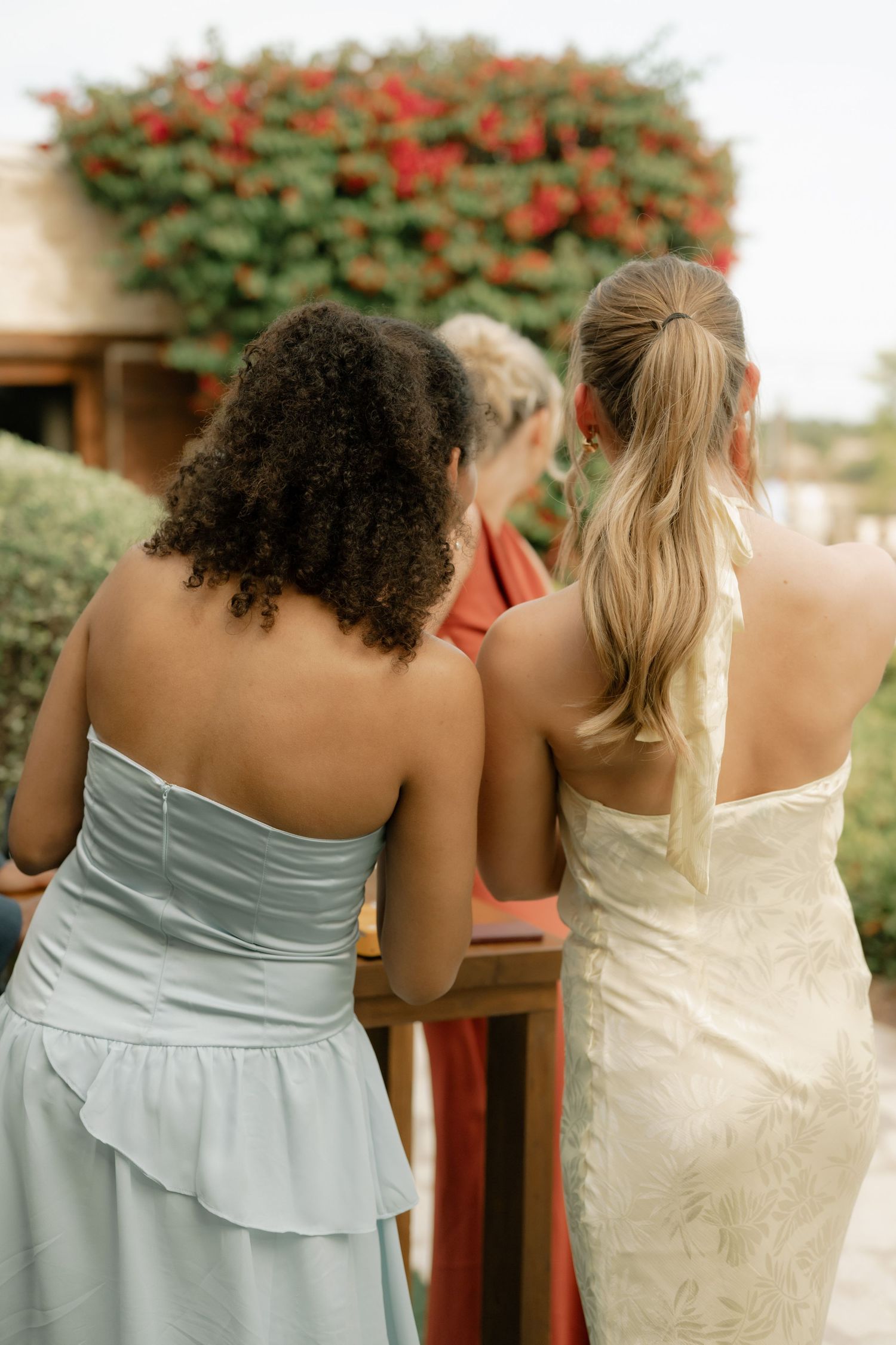 Two women in strapless dresses, one light blue and one white, stand together at an outdoor wedding ceremony.