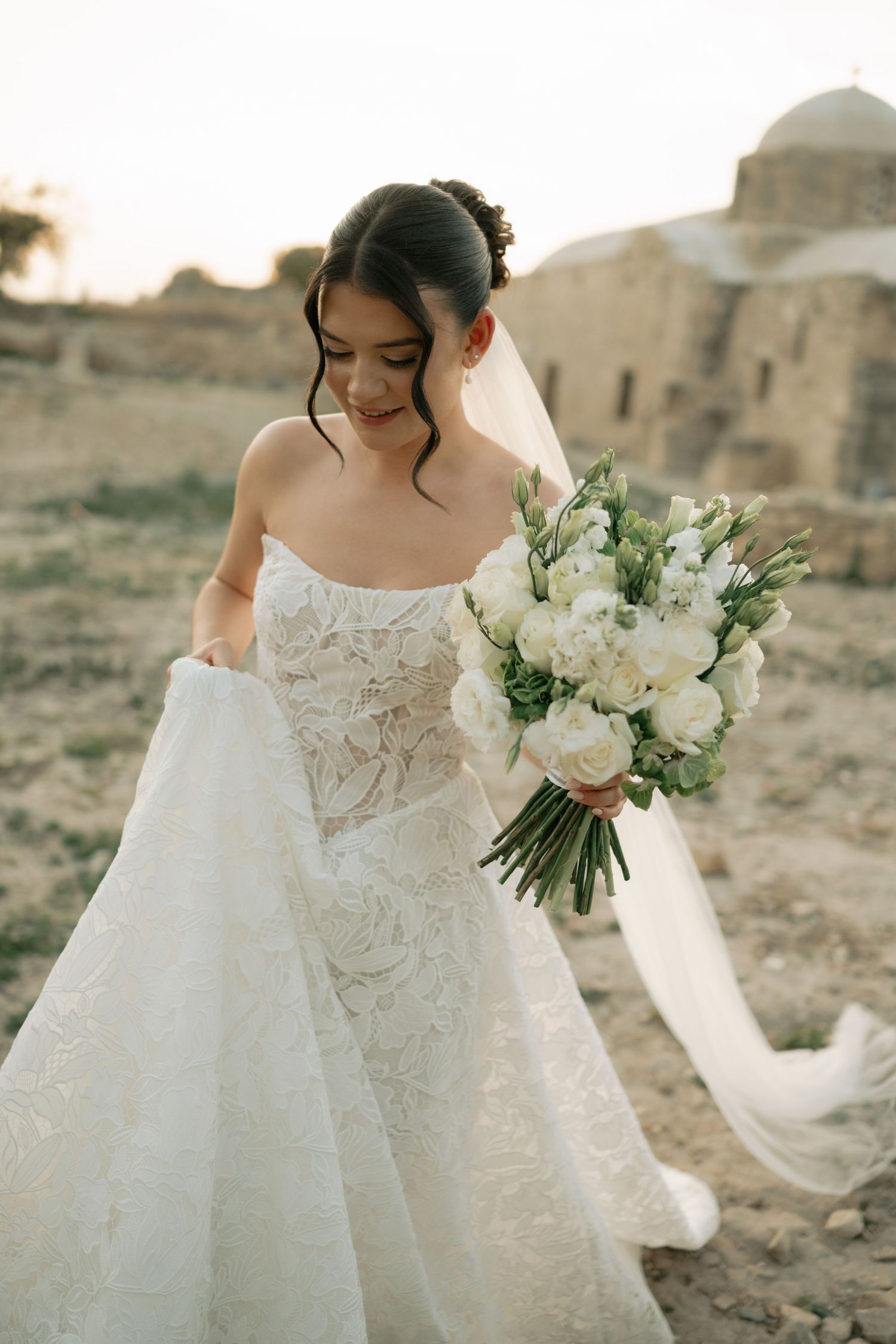 Bride in strapless lace wedding gown holding white floral bouquet, posing outdoors at golden hour.